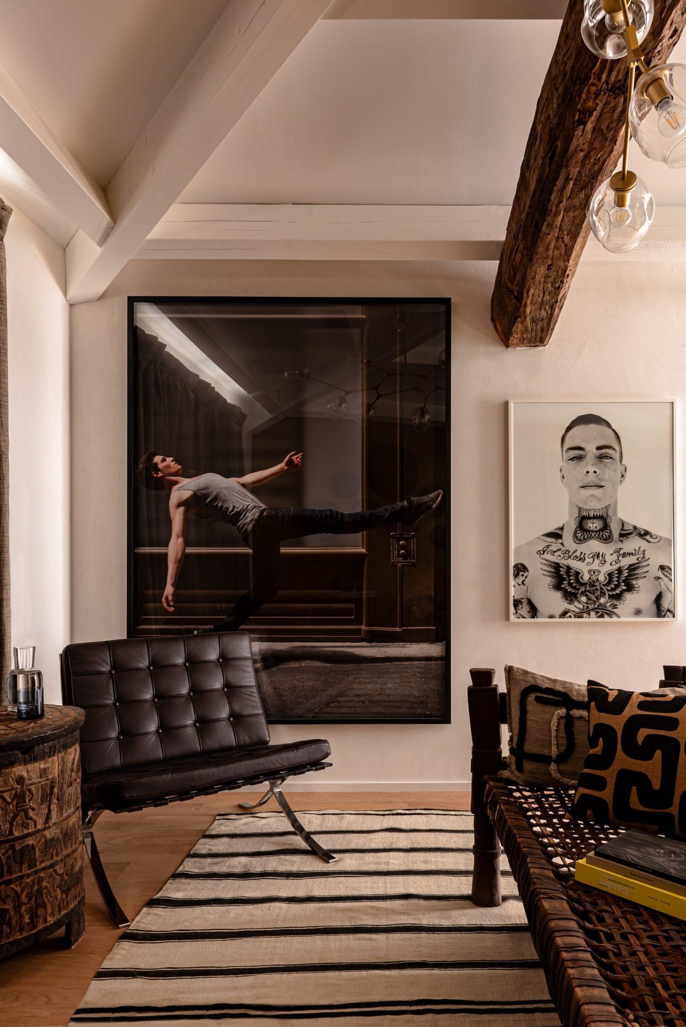 An elegant and modern living room features a large photograph of a floating silhouette on one wall and a monochrome portrait on another. The space includes a black leather chair, a wooden table, and patterned cushions on a bench. Exposed wooden beams and a striped rug add texture to the room.