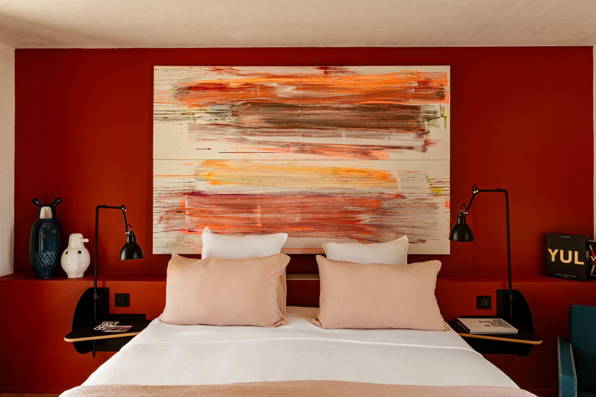 A modern bedroom features a double bed with beige and white bedding, flanked by bedside lamps. Above the bed hangs a large abstract painting in shades of red, orange, and beige. The room boasts bold red walls and decorative elements, including vases on a shelf behind the bed.