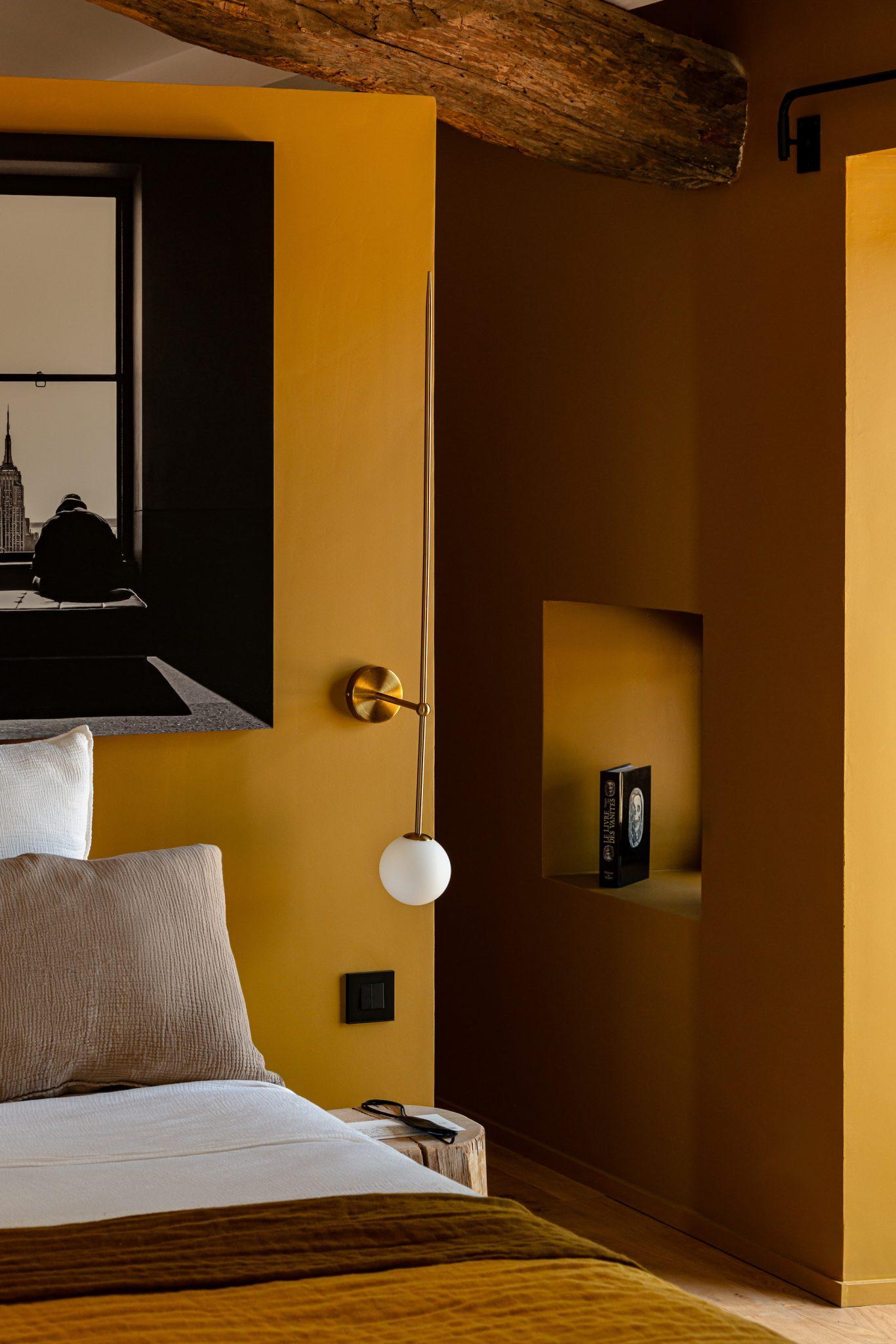 A cozy bedroom with mustard-yellow walls features a bed with white and brown pillows and blankets. A spherical, modern wall sconce hangs above the bed. A black-and-white photograph is hung on the wall, and a small alcove holds a single book.