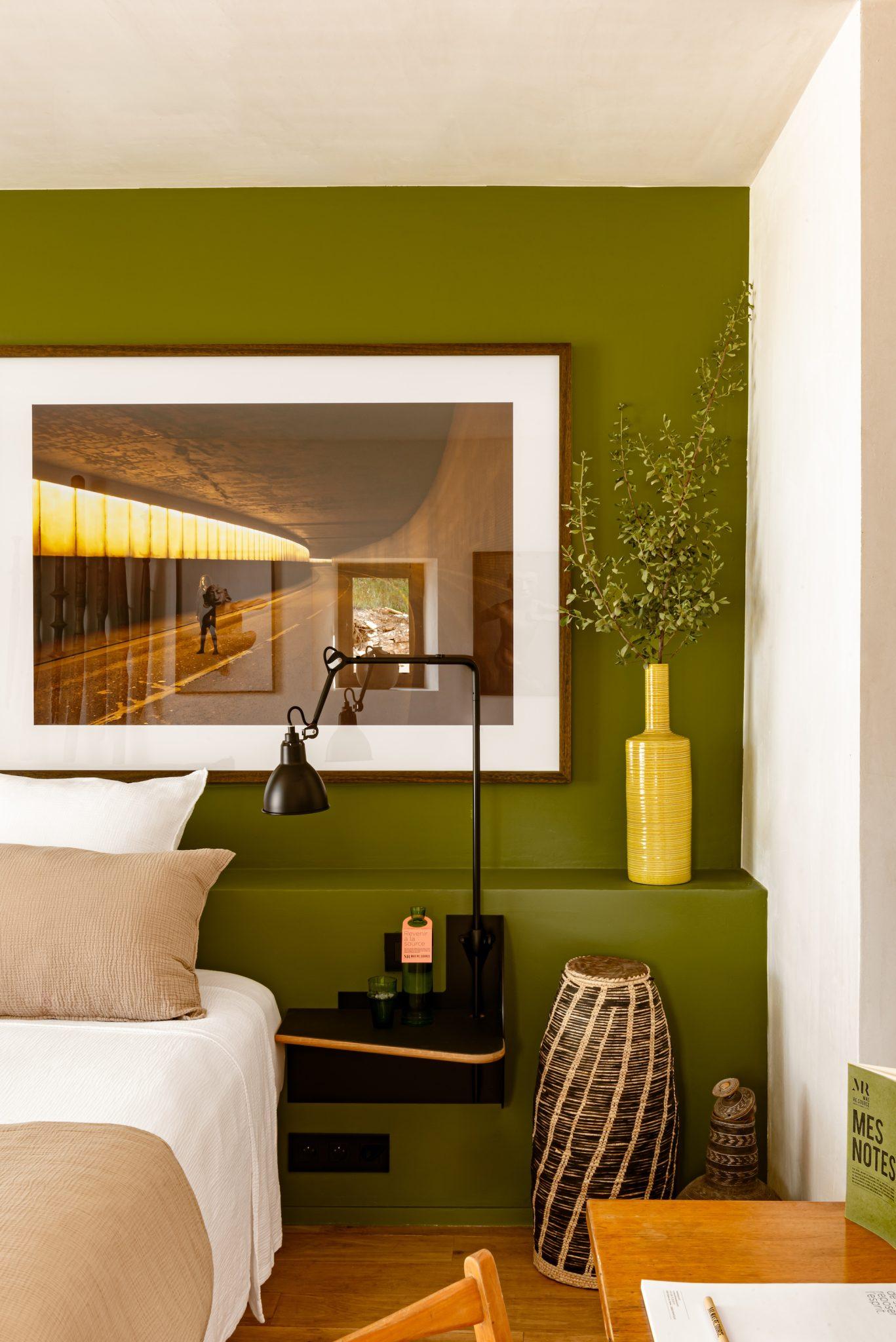 A cozy bedroom with an olive green accent wall features a framed photograph, a black reading lamp, and a yellow vase with green foliage. The bed is dressed in beige and white linens, and wooden decorations are placed on a small shelf and beside the bed.