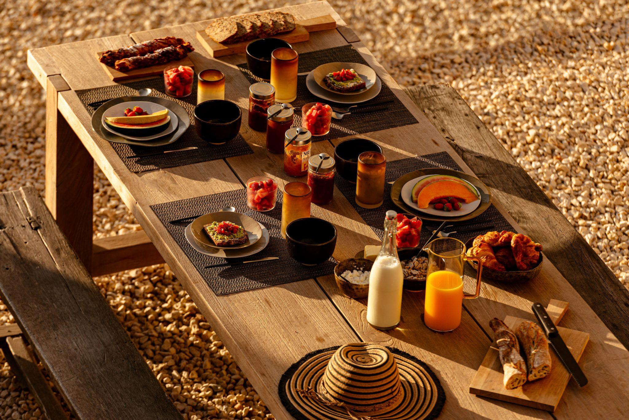 A wooden picnic table set for a meal features plates of food, glasses of orange juice and milk, and jars of preserves. The table is adorned with a straw hat, cut baguettes, and croissants. The setting is outdoors on a sunny day with a gravel floor.