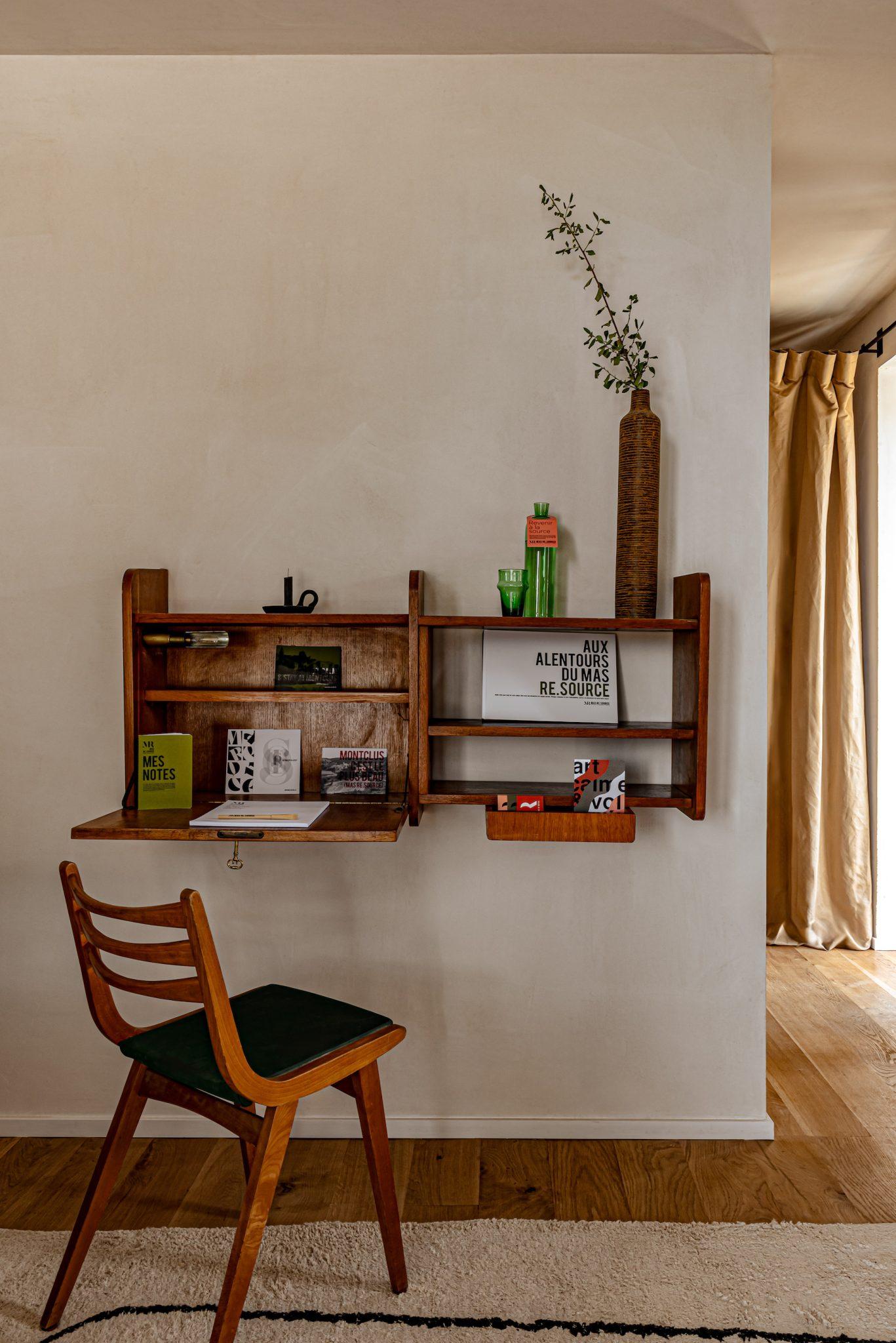 A comfortable and minimalist workspace features a wooden wall-mounted desk with shelves holding books, vases, and decorative objects. A wooden chair with a dark green seat cushion is placed in front of the desk. Light beige walls and a wooden parquet floor complete the scene.