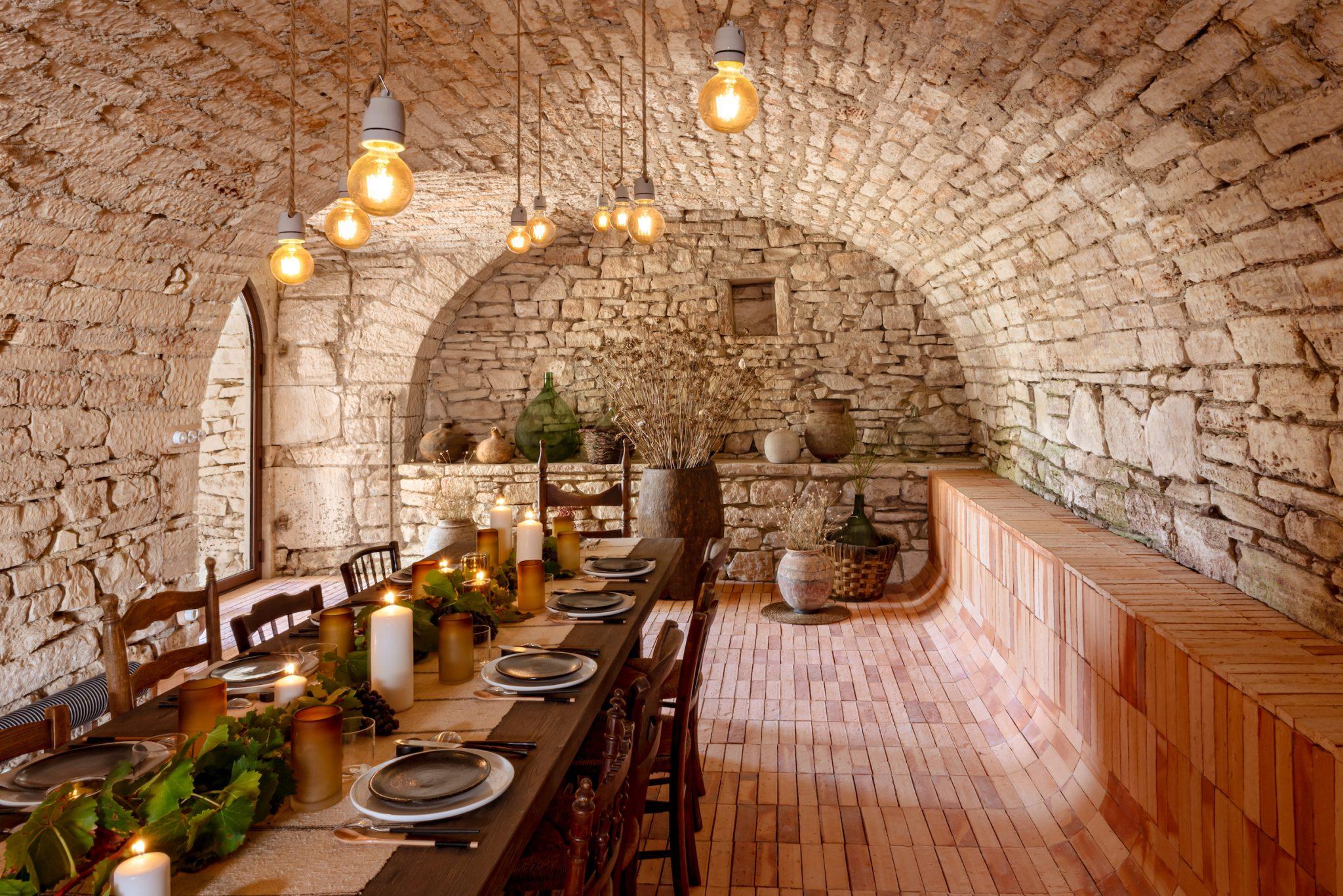 A rustic dining room with stone walls and a long wooden table set for a meal. The ceiling is vaulted, and several hanging lights are suspended above. The table is adorned with plates, glasses, plants, and candles. The lit bulbs enhance the warm and cozy atmosphere.