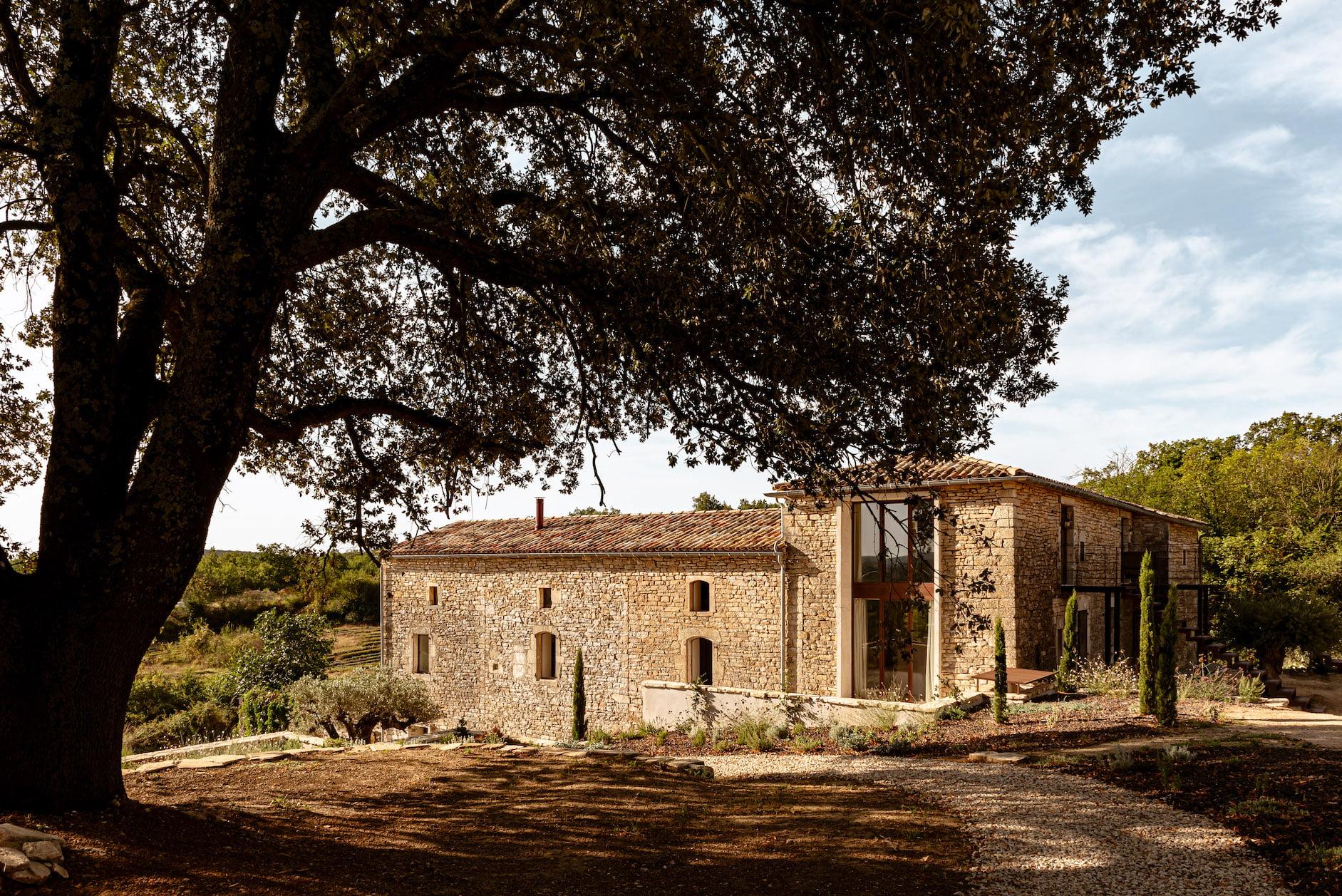 Une maison rustique en pierre avec de hautes fenêtres à cadre en bois, entourée de verdure et de grands arbres. Le chemin menant à l'entrée est ombragé par un grand arbre au premier plan. Le ciel est partiellement nuageux, projetant une lumière douce et naturelle sur le bâtiment.