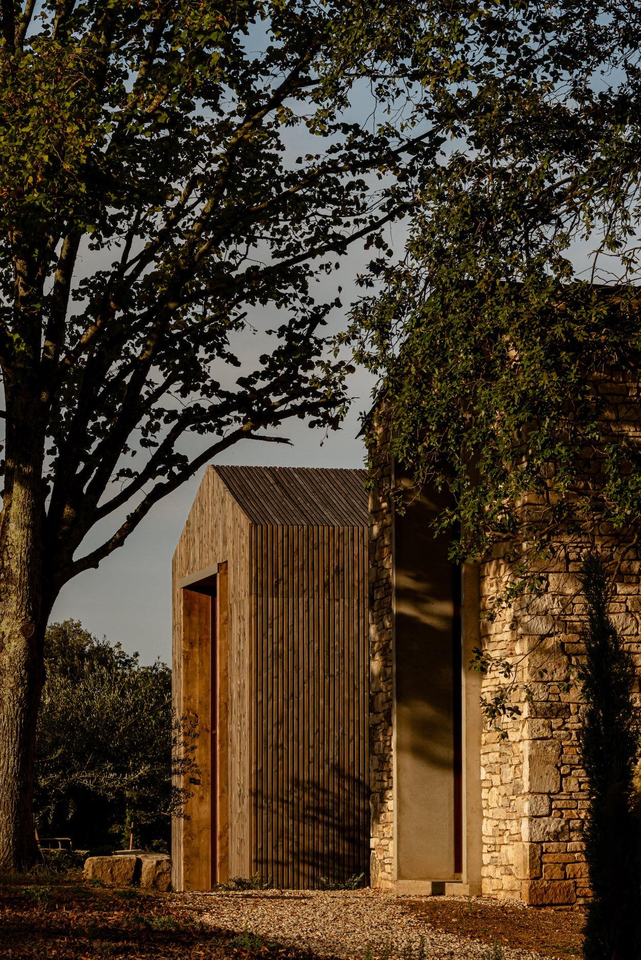 A rustic wooden structure, resembling a barn, stands amidst trees and greenery. Sunlight filters through the leaves, casting shadows on the weathered wooden planks and the adjacent stone wall. The scene exudes a serene and natural ambiance.