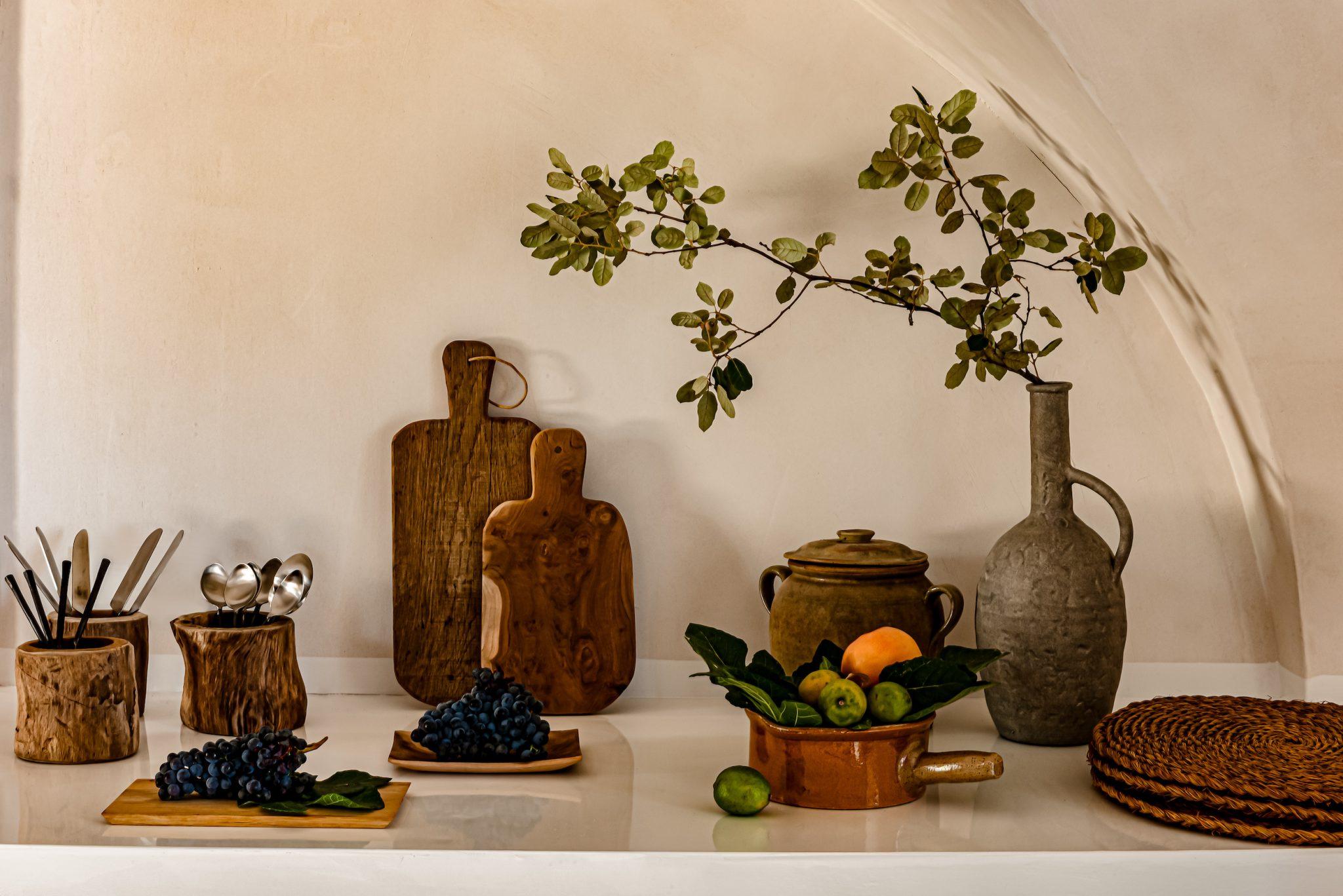 A kitchen countertop showcases a rustic décor with two wooden cutting boards, a few bunches of grapes, a ceramic vase filled with sprigs, a bowl of green fruits, and some utensils. Earthy tones and natural textures dominate the scene, creating a warm and inviting atmosphere.