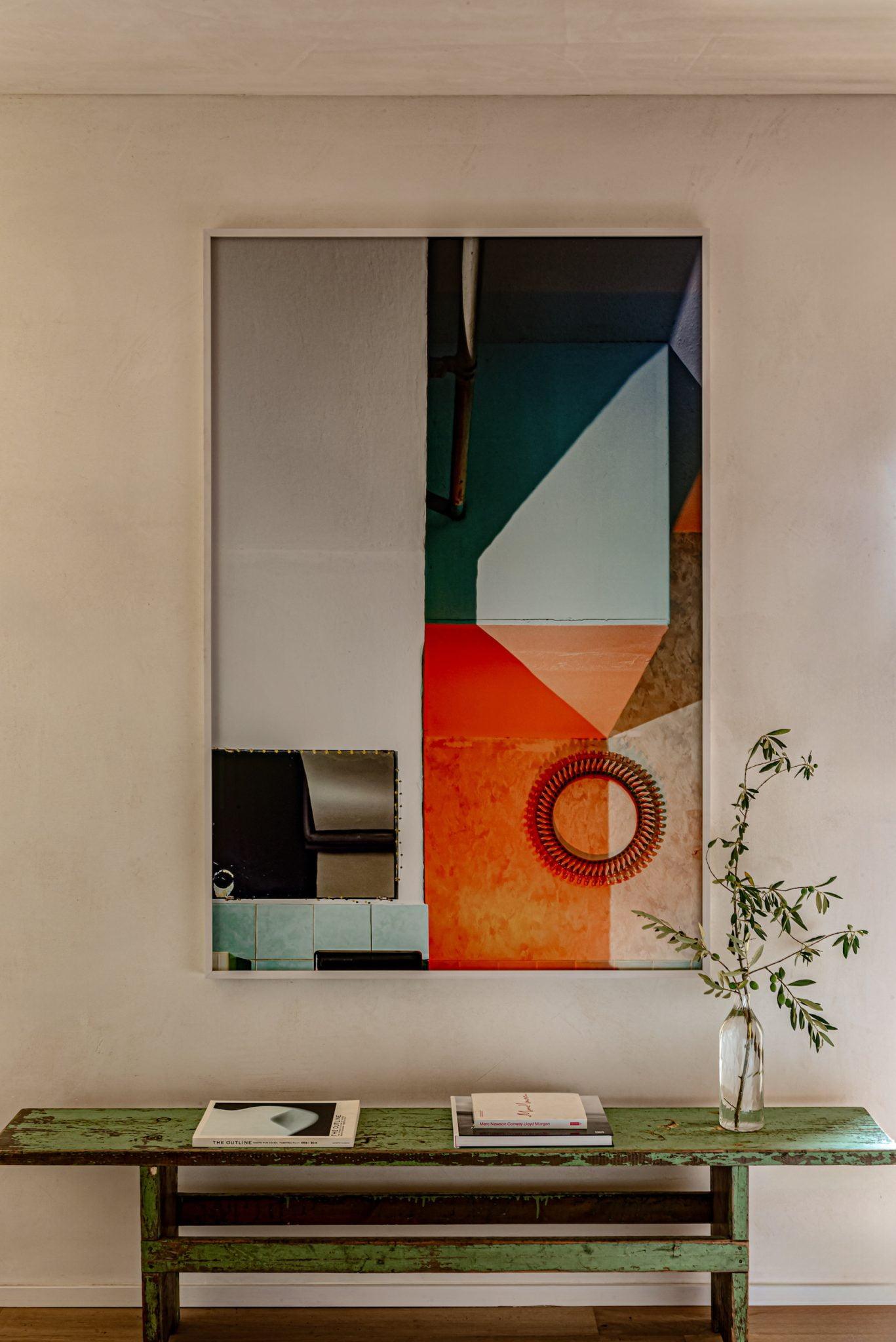 A framed abstract painting with geometric shapes and vibrant colors is hung on a cream-colored wall above a green wooden bench. A small tree branch in a glass vase and several books are placed on the bench, creating a minimalist and artistic décor.