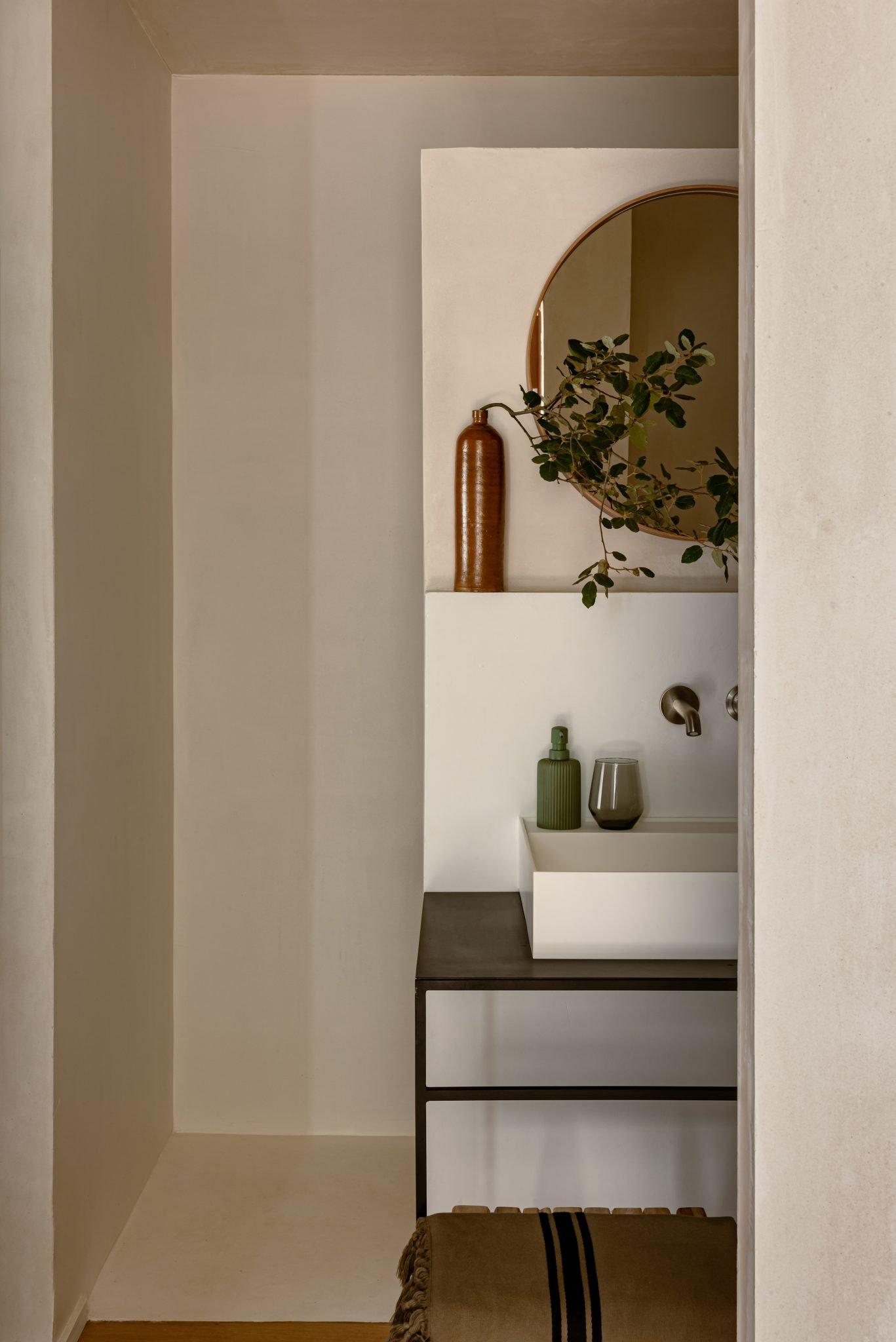 A minimalist bathroom features a white sink on a sleek black vanity. Above the sink, a circular mirror reflects the light, with a vase of greenery and a bottle on a shelf. Neutral tones dominate the space, creating a calm and modern aesthetic.