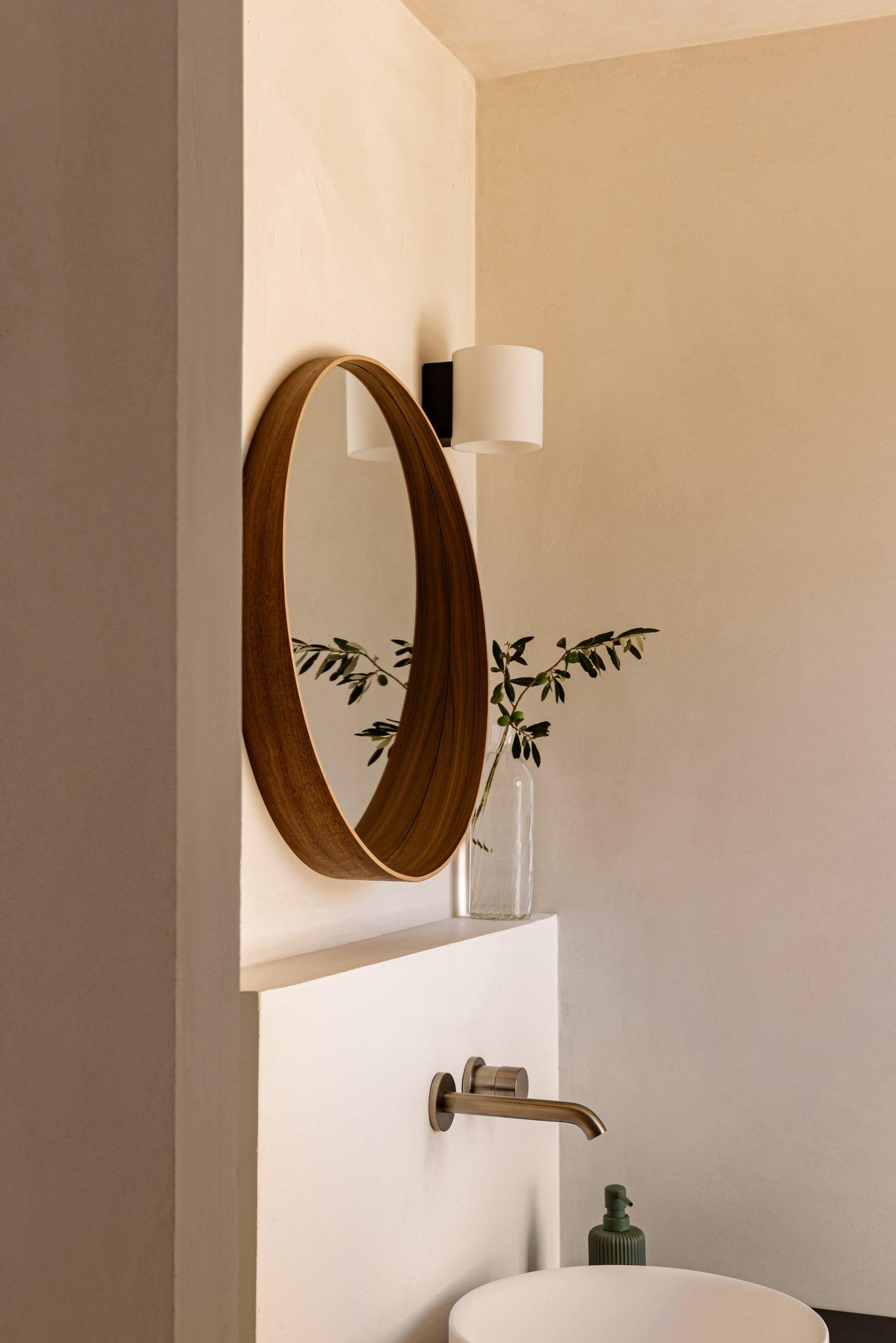 A minimalist bathroom features a round wooden-framed mirror mounted on a smooth beige wall. Below, a small green potted plant in a glass container is placed next to a round white sink with an elegant faucet. A wall light with a white lampshade is positioned above the mirror.