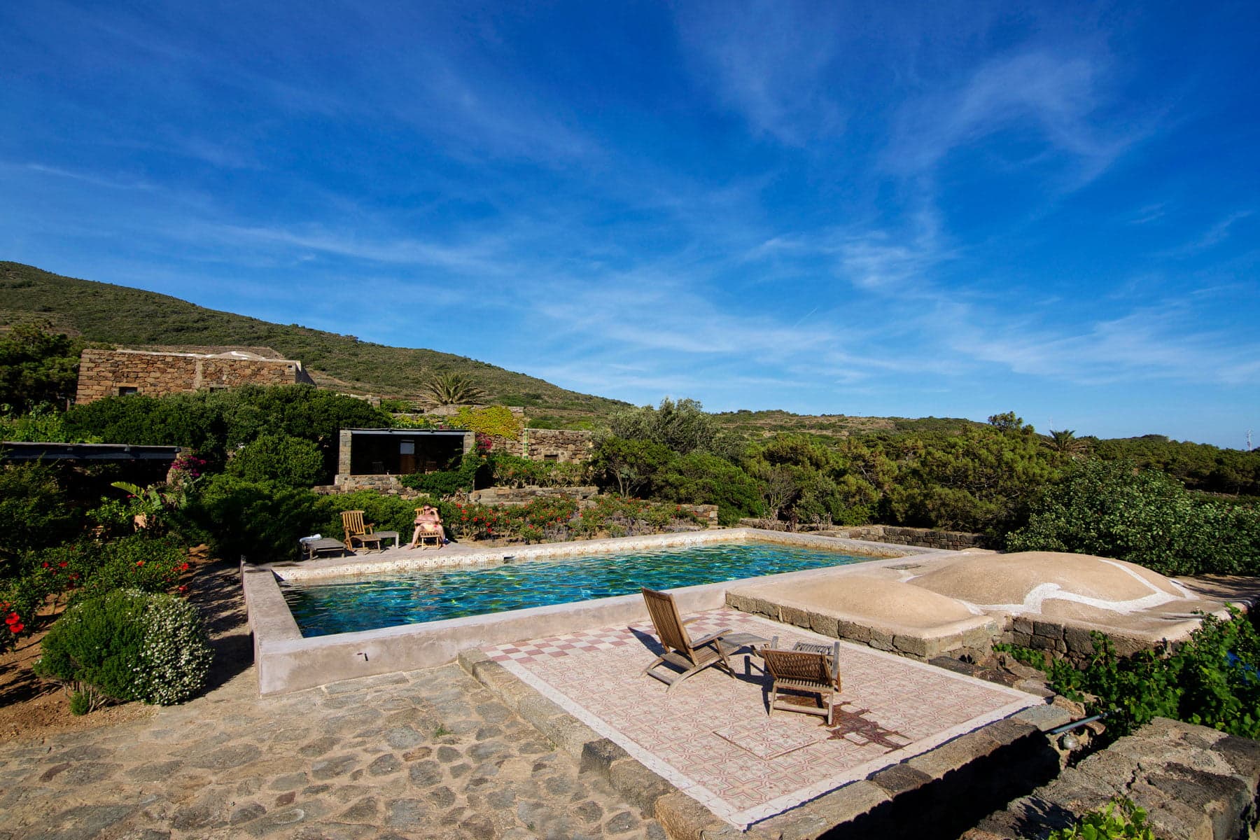 Vue panoramique d'une piscine dans un espace extérieur isolé avec des chaises longues autour. La piscine est entourée d'une végétation luxuriante et d'un patio en pierre rustique. En arrière-plan, il y a un terrain montagneux sous un ciel bleu clair. Une personne est assise près de la piscine.