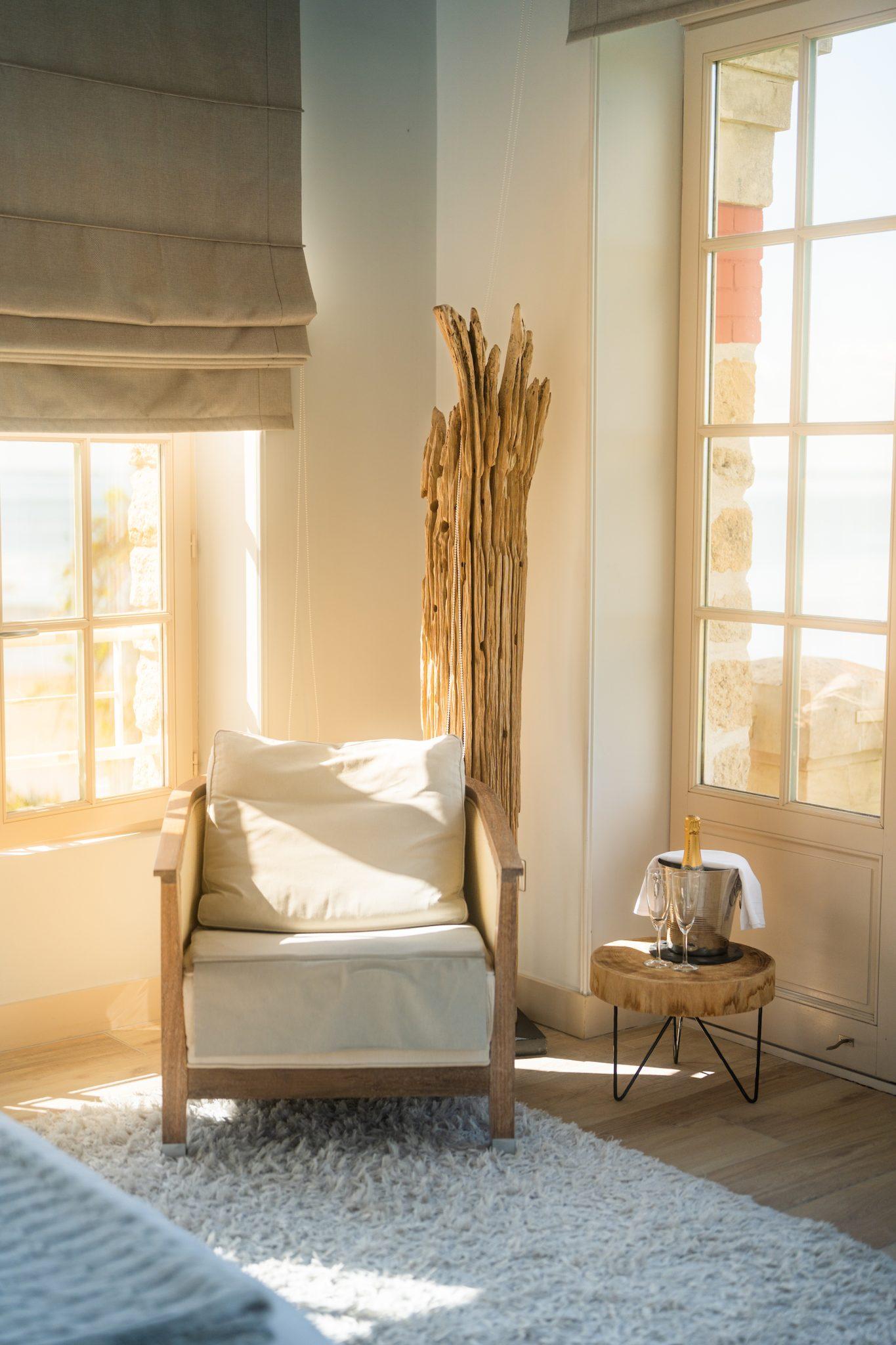 A comfortable, sunny living space with an upholstered wooden armchair on a soft white carpet. Next to the armchair is a small round wooden table with a bucket containing a bottle of champagne and two flutes. A floor lamp made from driftwood stands by the large windows.