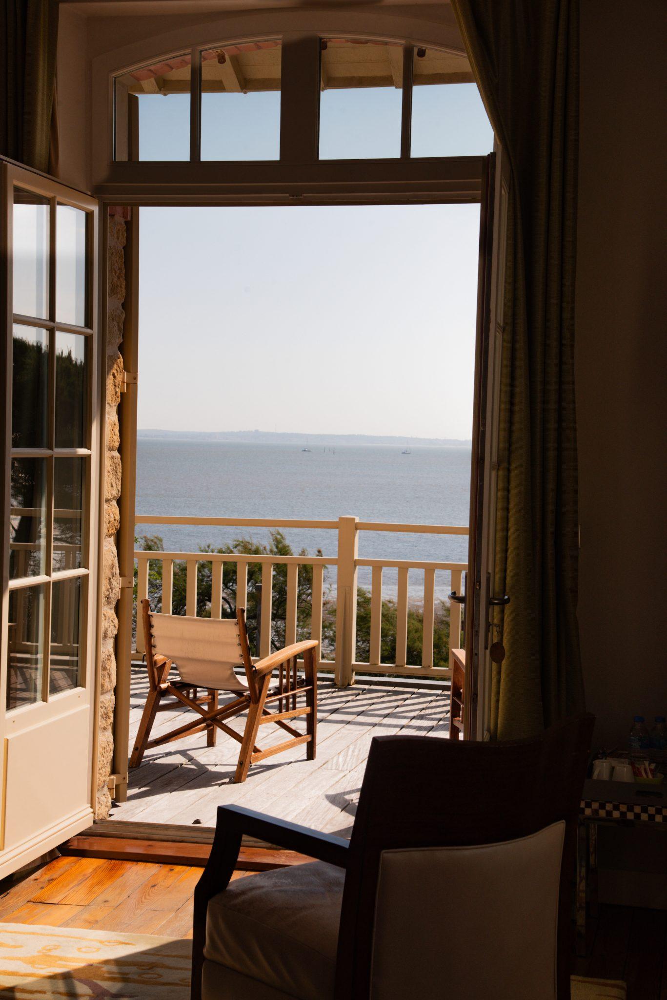 A room with an open door leading to a balcony overlooking a serene body of water. The balcony has a wooden chair and is surrounded by railings. Sunlight streams into the room through large windows, creating a warm and inviting atmosphere.