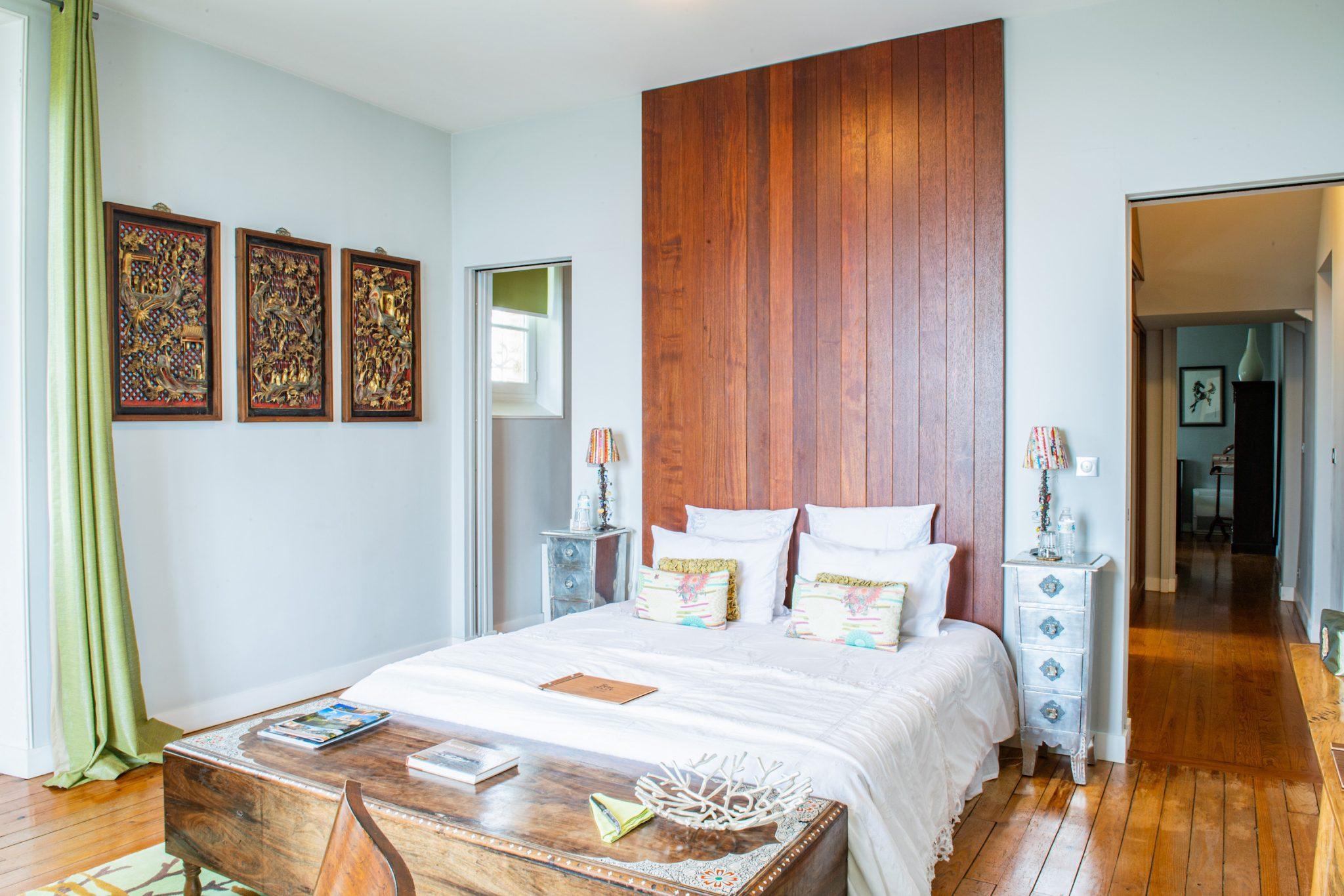 Une chambre confortable avec une grande tête de lit en bois, un lit avec des draps blancs et des oreillers colorés. Deux tables de nuit argentées avec des lampes se trouvent de chaque côté du lit. Il y a trois œuvres d'art décoratives encadrées sur le mur et un plancher en bois accentué par un banc en bois au pied du lit.