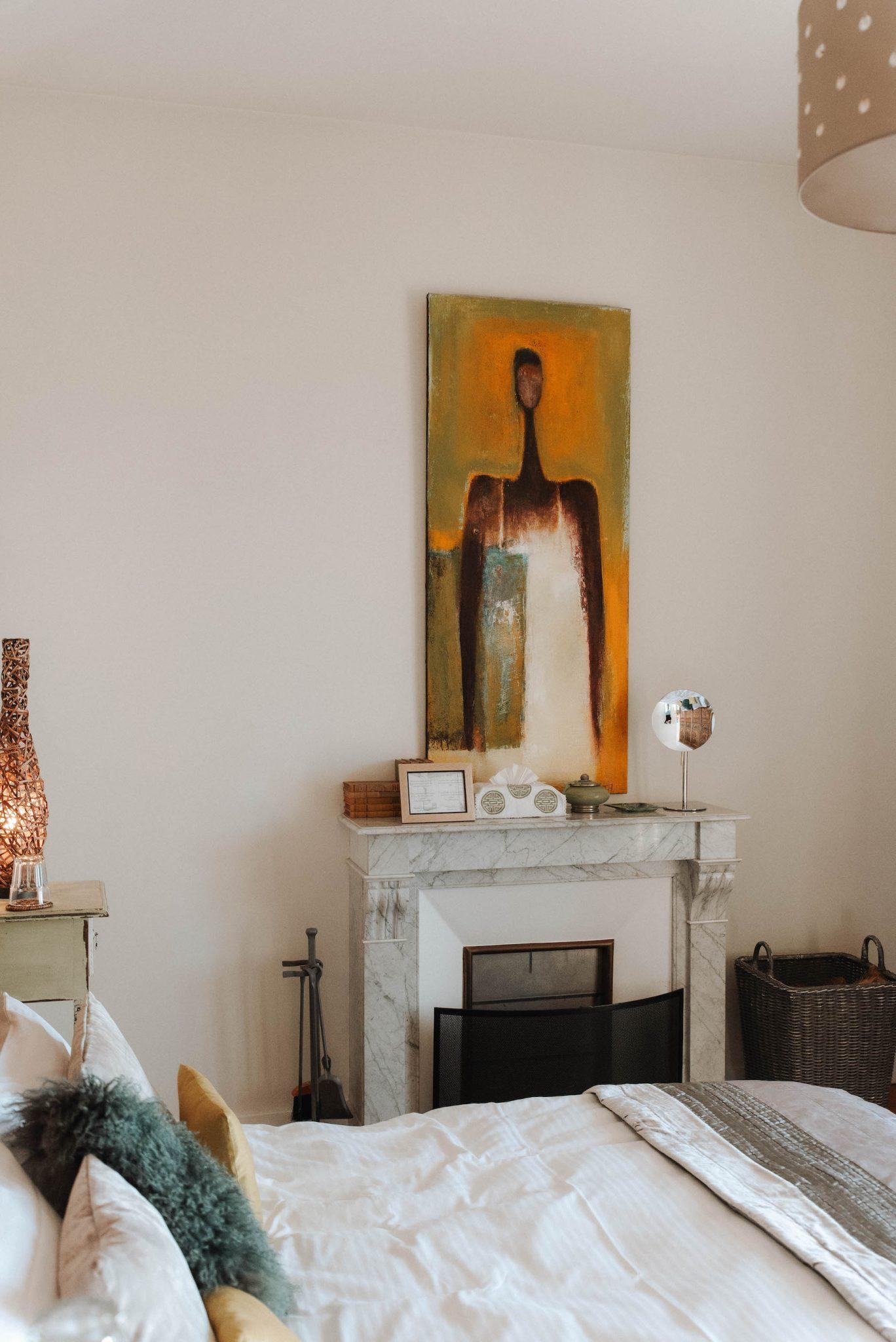 A cozy room features a white marble fireplace with a modern abstract painting hung above it. To the left, there is a bedside table with a lamp, and to the right, a wicker basket is placed. In the foreground, the bed is adorned with decorative pillows and a blanket.