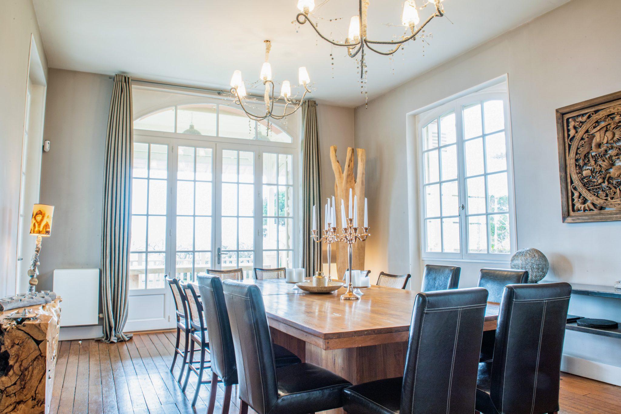 Une salle à manger spacieuse avec une grande table en bois entourée de chaises en cuir noir. Deux lustres sont suspendus au plafond et de hautes fenêtres avec des rideaux laissent entrer la lumière naturelle. Une œuvre d'art décorative en bois orne le mur et un candélabre est posé sur la table.