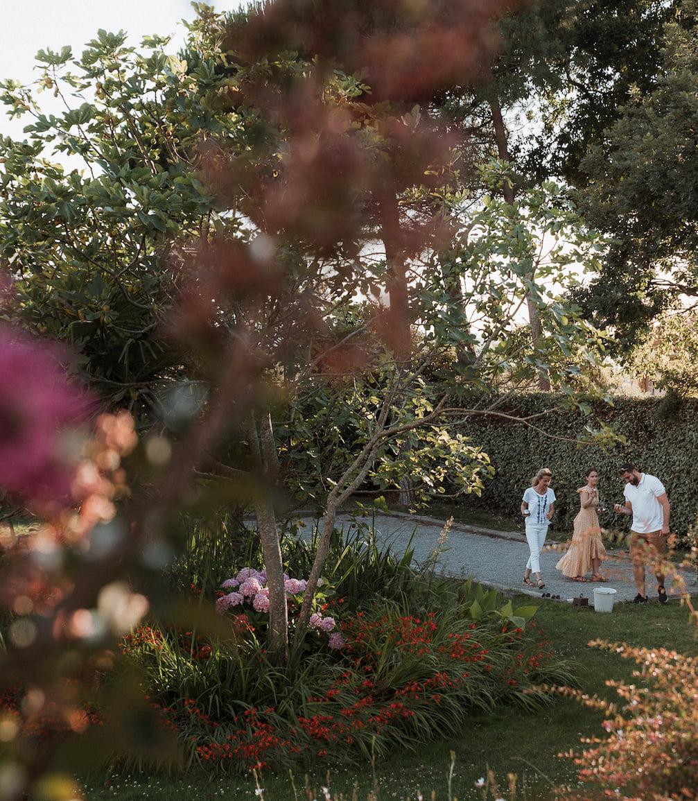 Three people stand at the edge of a gravel path in a lush garden. The foreground features vibrant flowers and dense foliage, partially obscuring the view. Tall trees and additional greenery form the background, creating a serene and natural setting.