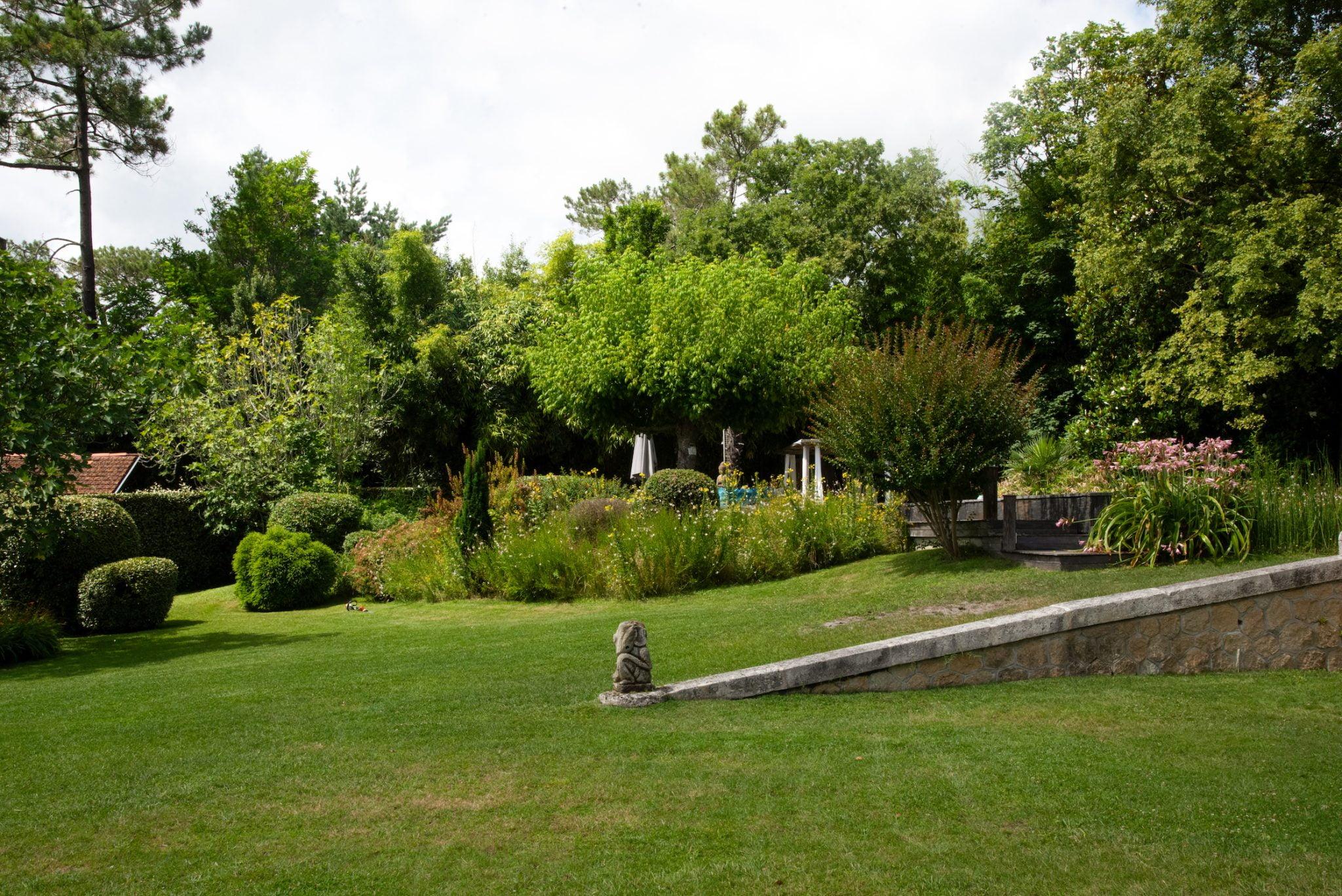 A lush garden with a variety of trees, shrubs and flowers. In the centre, there is a shady seating area under a large tree. A stone path and low stone wall border the well-kept lawn. The background features dense vegetation, suggesting a serene, peaceful outdoor space.