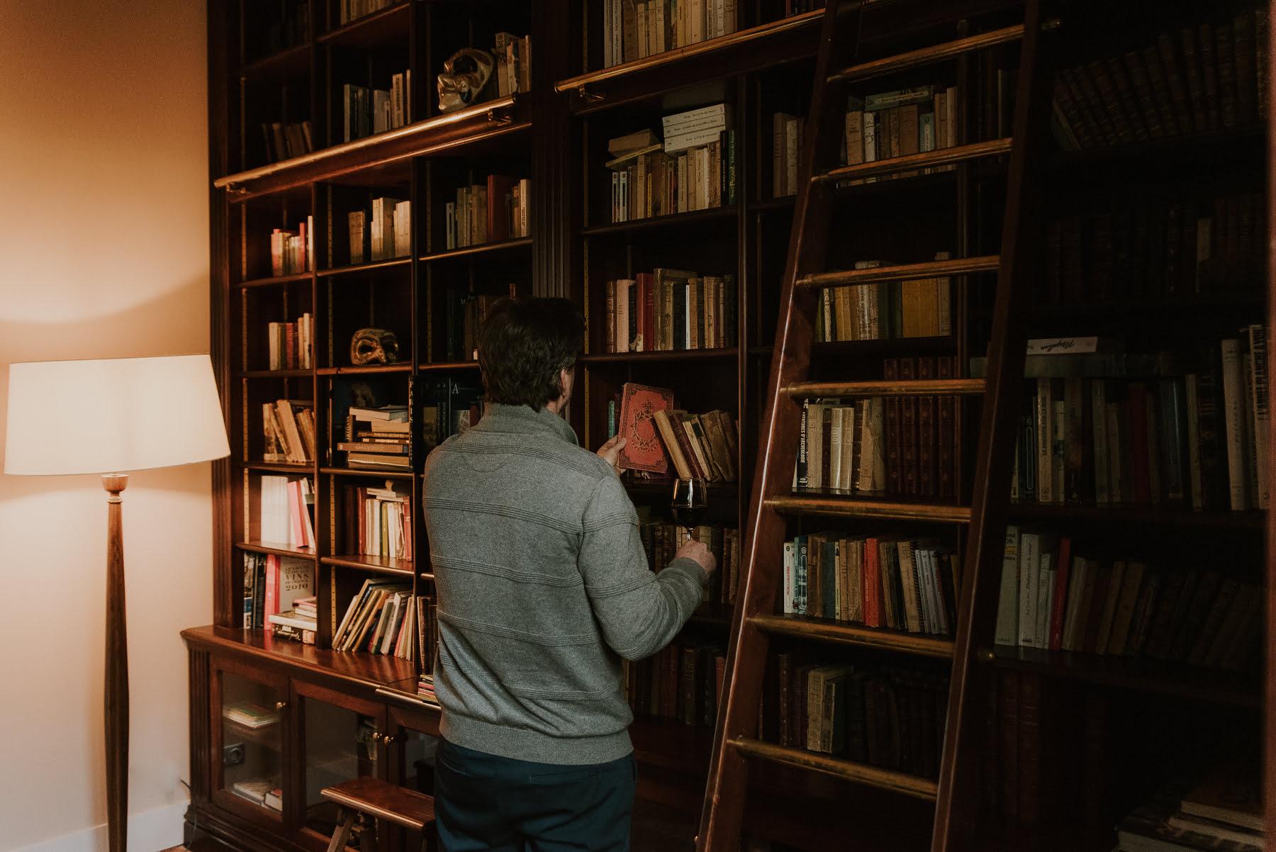 A person in a grey jumper chooses a book from a large wooden shelf filled with books. A wooden ladder is leaning against the shelf and a floor lamp is lit nearby, casting a warm glow over this cosy library-like room.