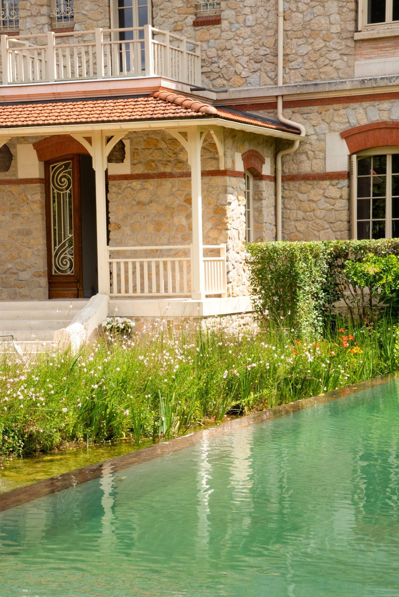 A charming stone house with a porch and wooden door can be seen beside a serene pond. The turquoise water of the pond reflects the image of the house. Around the pond are lush green shrubs and tall grasses with white and orange flowers.