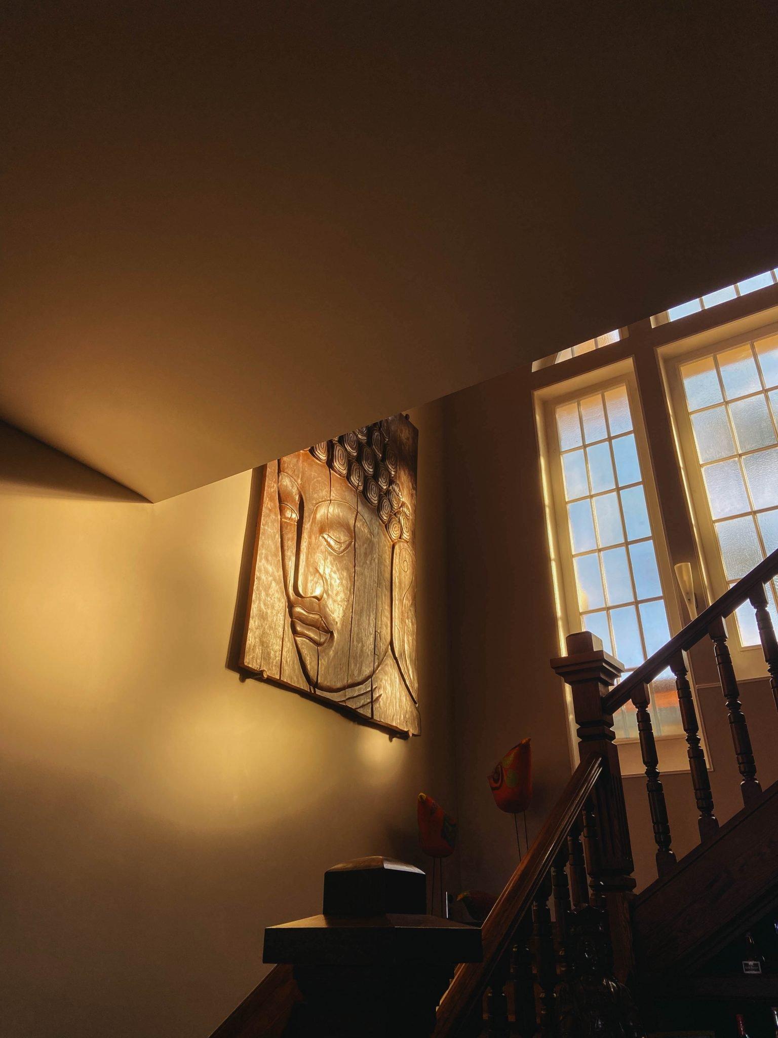 A staircase with a wooden handrail is bathed in the warm sunlight streaming through large windows. A prominent piece of artwork depicting a serene Buddha face is hung on the wall. At the foot of the staircase, a small stack of objects is visible, adding a touch of cozy clutter.