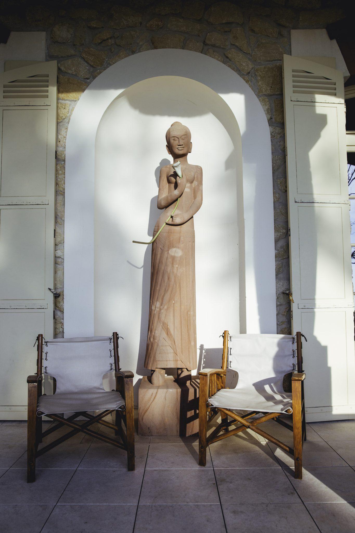 A tall, slender statue of a figure with a serene face stands in an arched niche against a stone wall. Two wooden chairs with white fabric seats are placed on either side of the statue, creating a symmetrical configuration on a tiled floor.