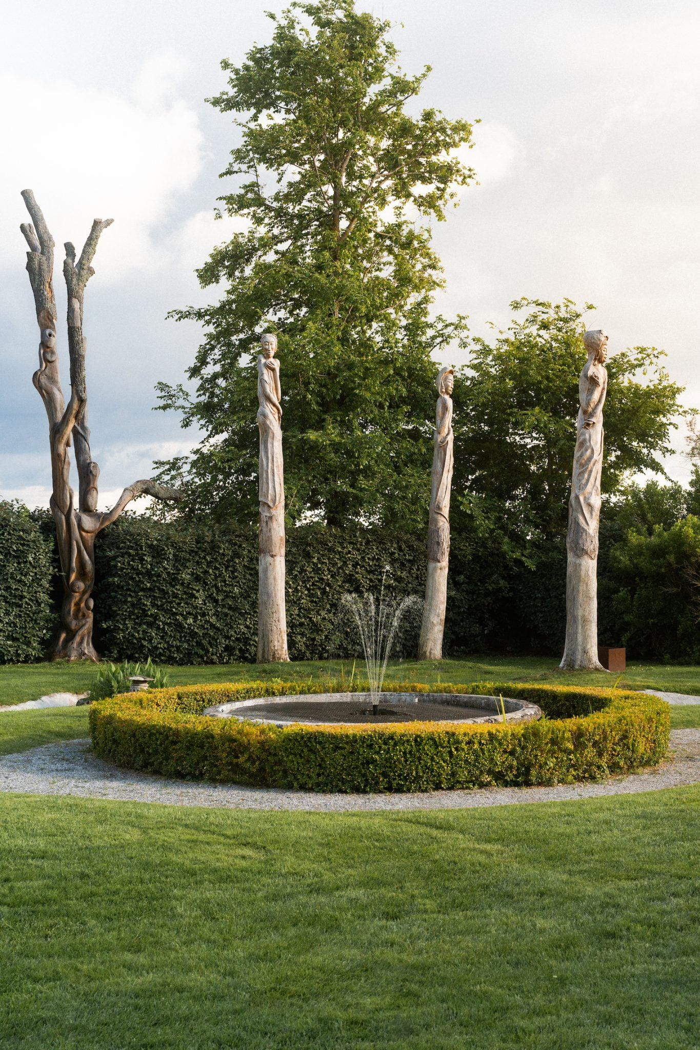 A circular garden fountain is surrounded by a low green hedge and a gravel path. Four rustic wooden sculptures resembling large trees are arranged around the fountain. In the background, lush green hedges and a large tree enhance the serene garden setting.