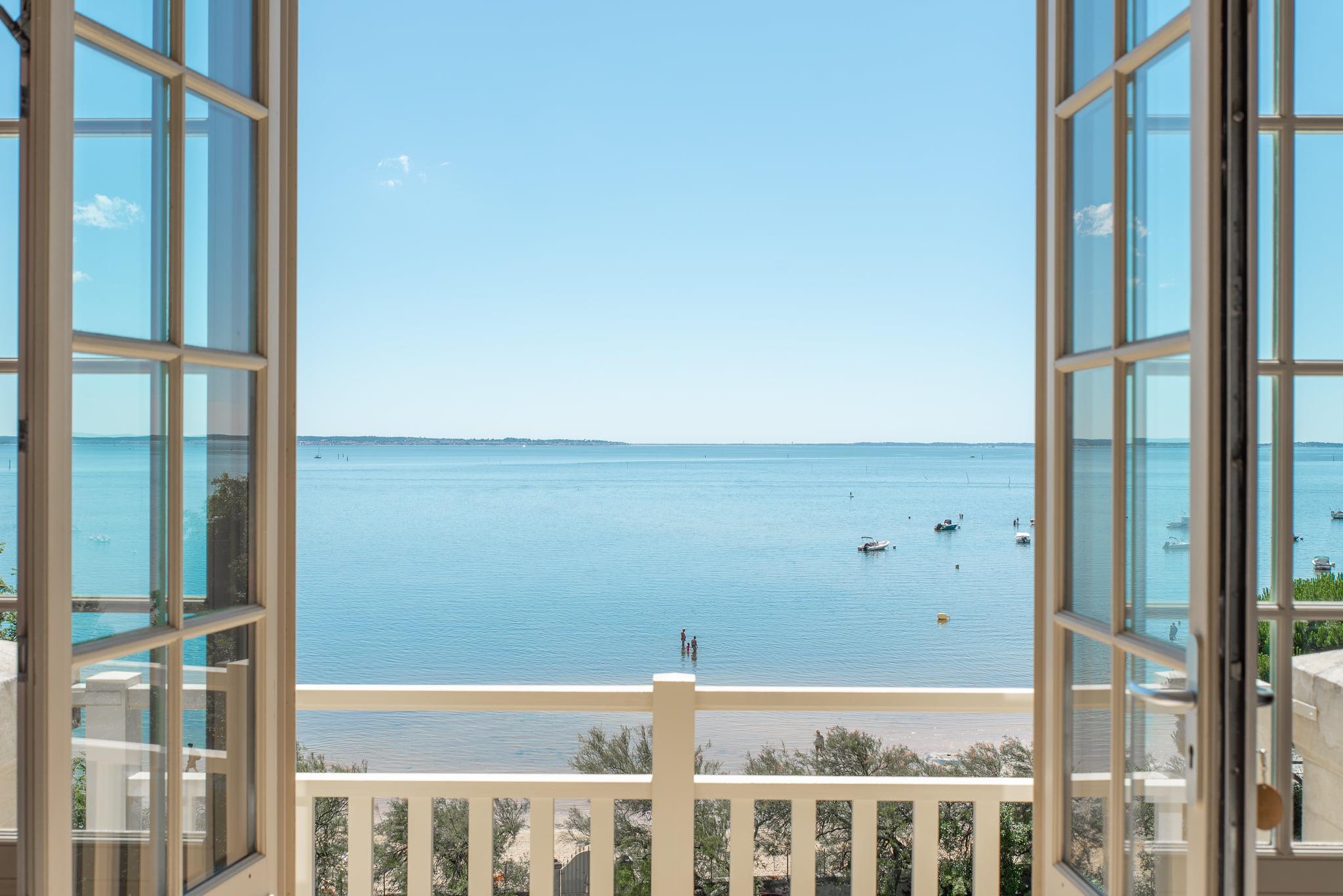 Vue panoramique à travers les portes-fenêtres ouvertes de la terrasse révélant un océan bleu calme, un ciel clair et un horizon lointain. Une petite plage de sable avec quelques bateaux ancrés près du rivage est visible, encadrée par la balustrade du balcon. Une végétation clairsemée est visible sur les bords de l'image.