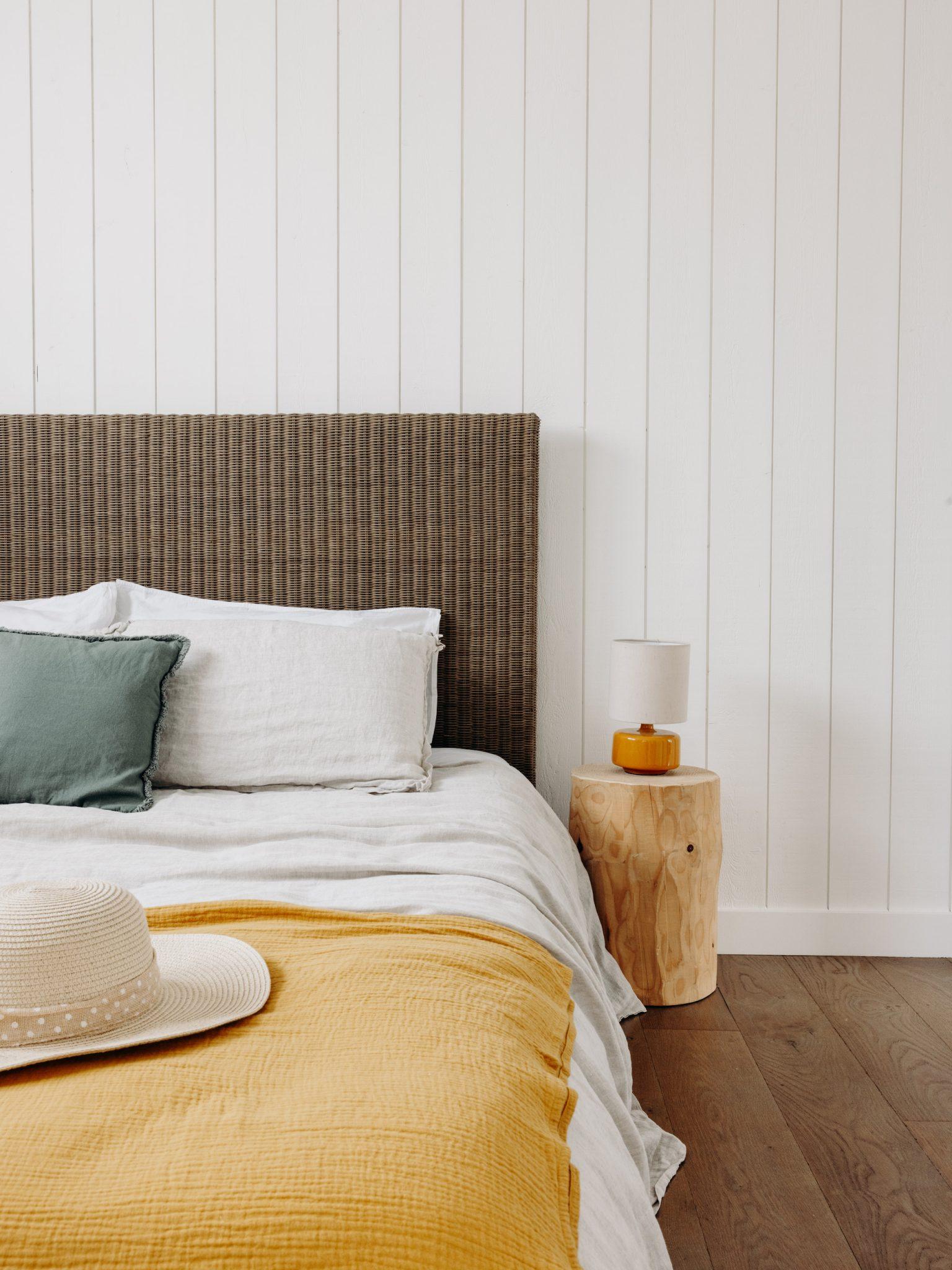 A cozy bedroom with a light-paneled wall, a brown upholstered headboard, and a bed dressed in white and gray bedding with green and white pillows. A yellow blanket covers the foot of the bed, with a straw hat resting on top. A wooden nightstand holds a lamp and a small decorative item.