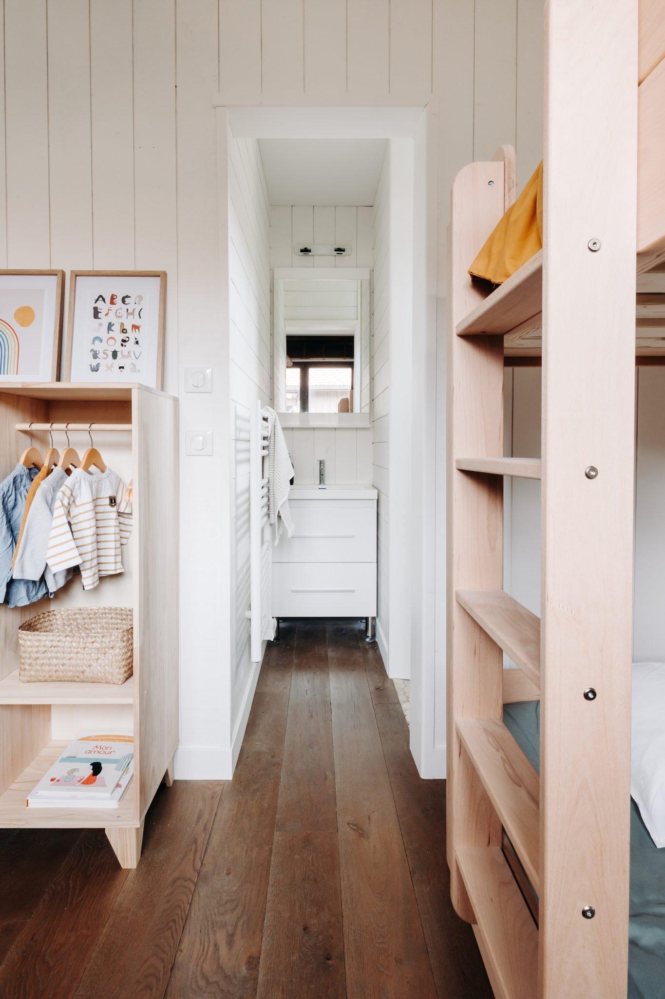 A cozy small children's room features wooden bunk beds, a wardrobe with neatly hung clothes, and a wicker basket. The floor is made of dark wood, and a narrow hallway leads to a small white bathroom with a vanity and mirror. Pastel wall art decorates the space.