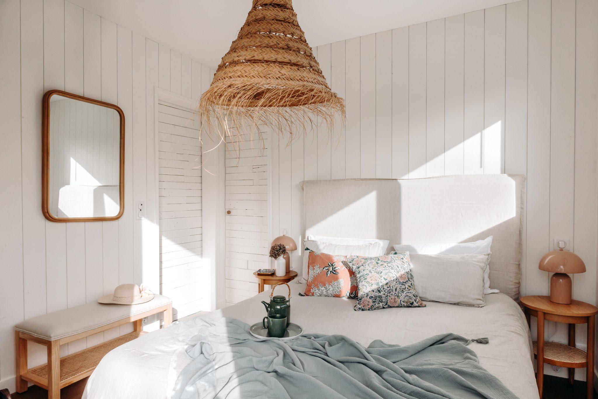 A cozy bedroom with white shiplap walls, a large woven pendant light, and a light-colored bed adorned with several pillows and a throw blanket. There’s a bench with a hat on it, a mirror on the wall, and two nightstands with lamps and decorative items.