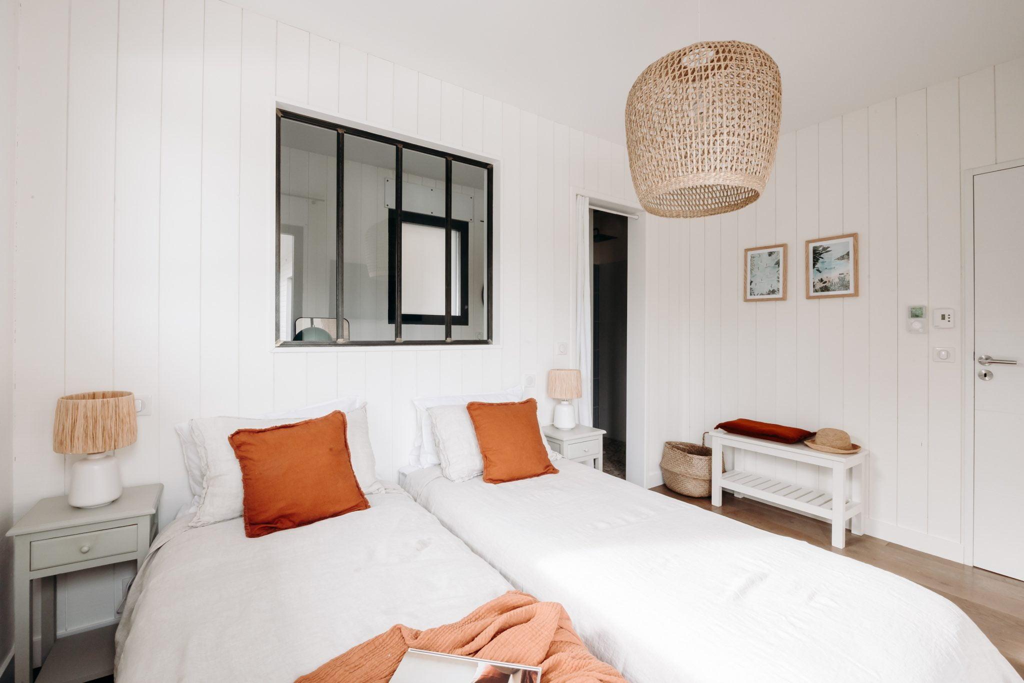 A cozy bedroom with twin beds dressed in white sheets and orange pillows. Each bed has a light green nightstand with a lamp. A woven pendant light hangs from the ceiling. The room features white paneled walls, framed pictures, and a small bench with baskets.