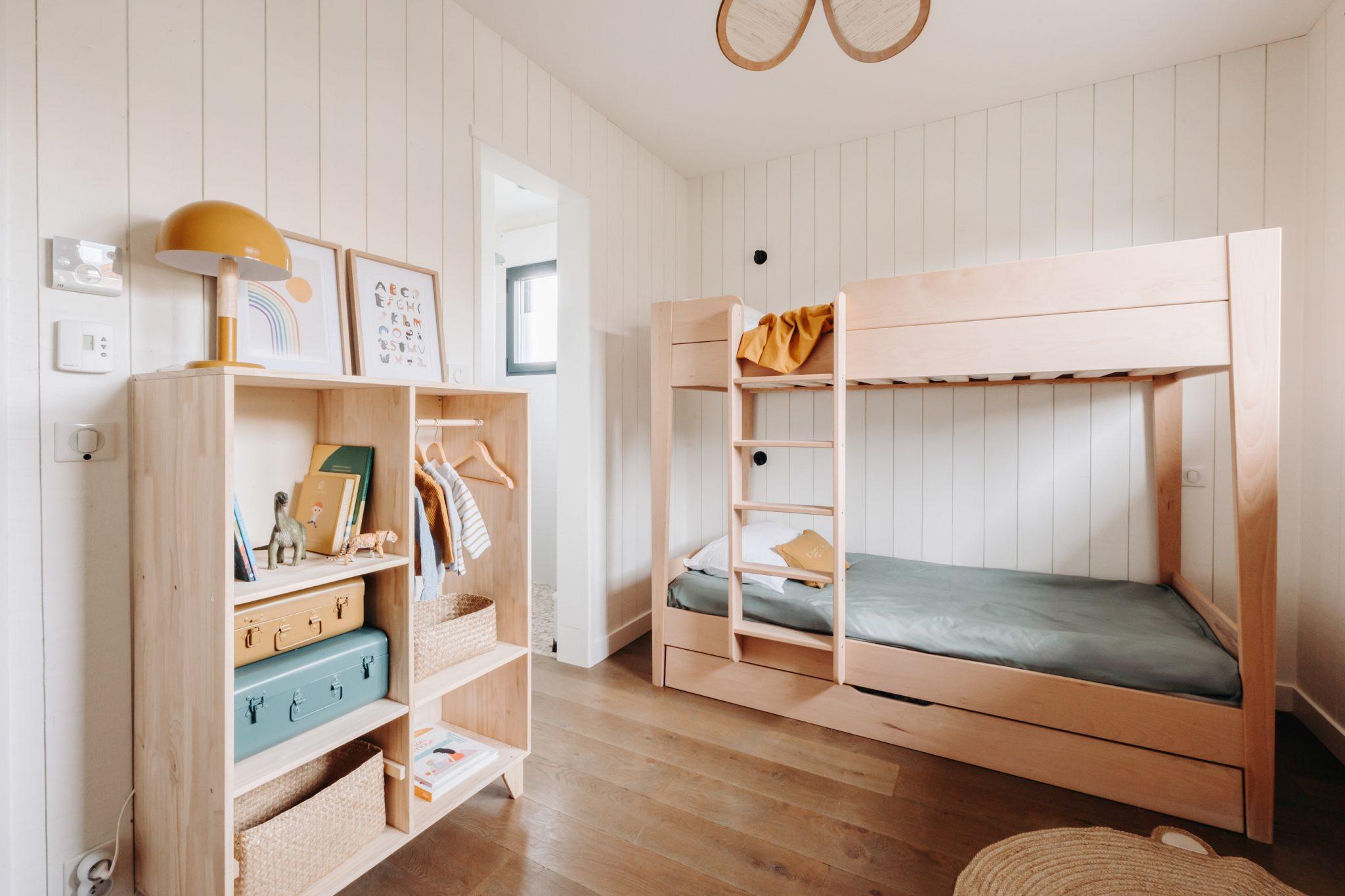 A cozy and minimalist children's room with white paneled walls and hardwood floors. It includes a wooden bunk bed with green bedding, a wooden shelf filled with toys, books, and clothes, a mustard-colored lamp, and a framed rainbow print on the wall.