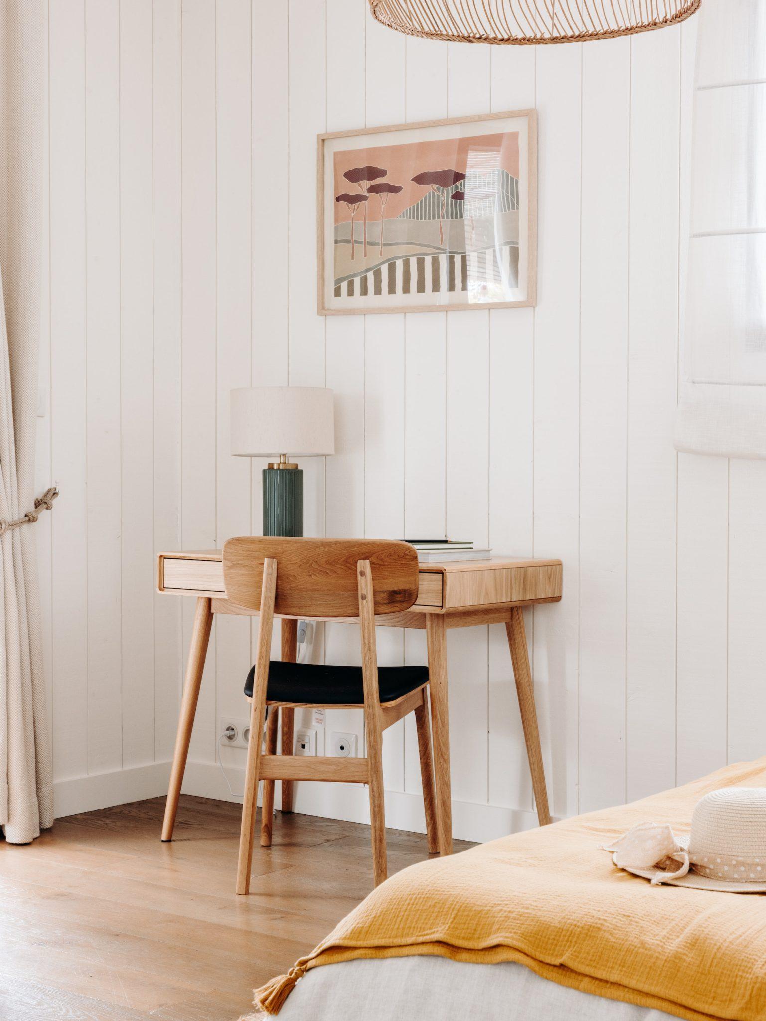 A minimalist workspace featuring a wooden desk with a matching chair. The desk has a lamp, books, and decorative items. Above it hangs a framed piece of art. The room has white wooden walls, a window with sheer curtains, and a bed with a yellow blanket in the foreground.