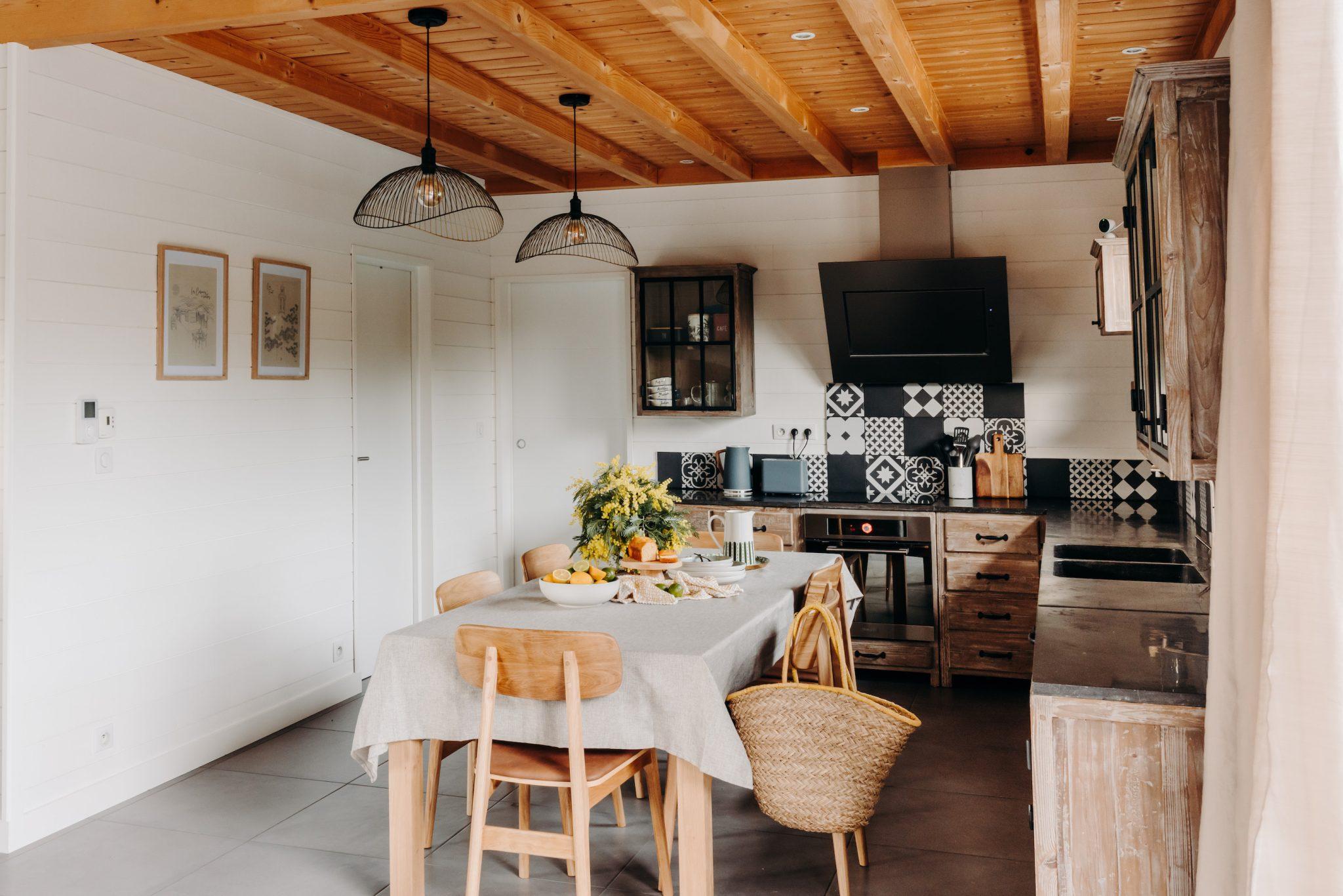 A cozy kitchen with a wooden ceiling and beams, white walls, and a black and white tiled backsplash. It includes a dining table covered with a gray tablecloth, surrounded by wooden chairs. The kitchen is decorated with potted plants and framed pictures.