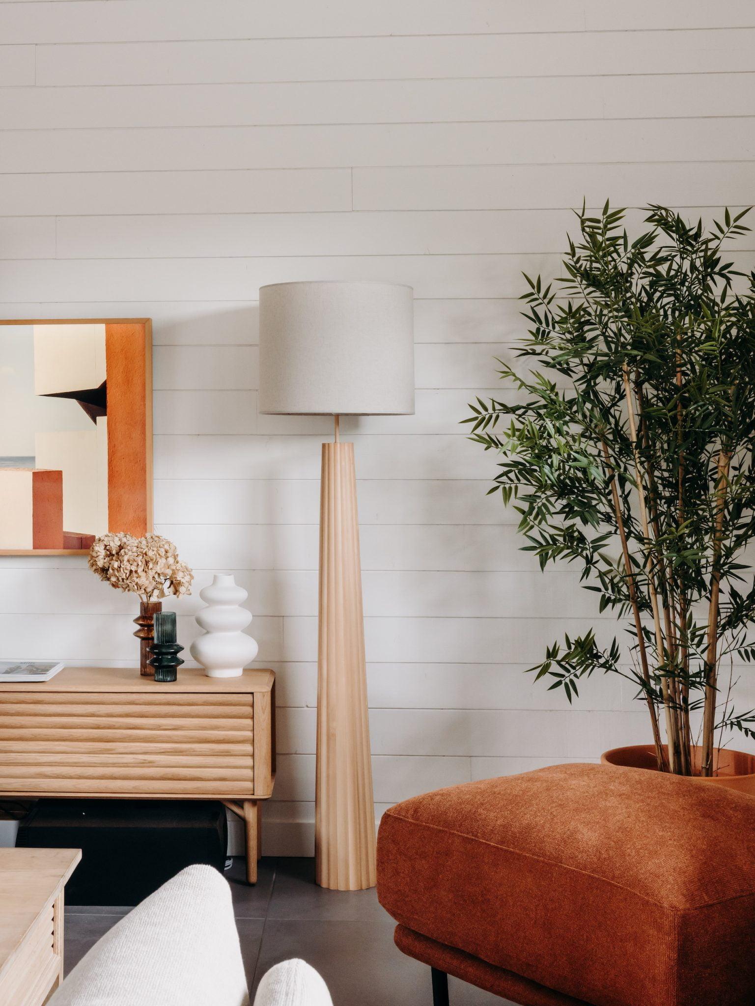 A cozy living room features a large lamp with a wooden base, a potted plant, a wooden sideboard with a decorative vase, and a modern painting on the wall. An orange armchair is placed in the foreground, adding warmth to the space.