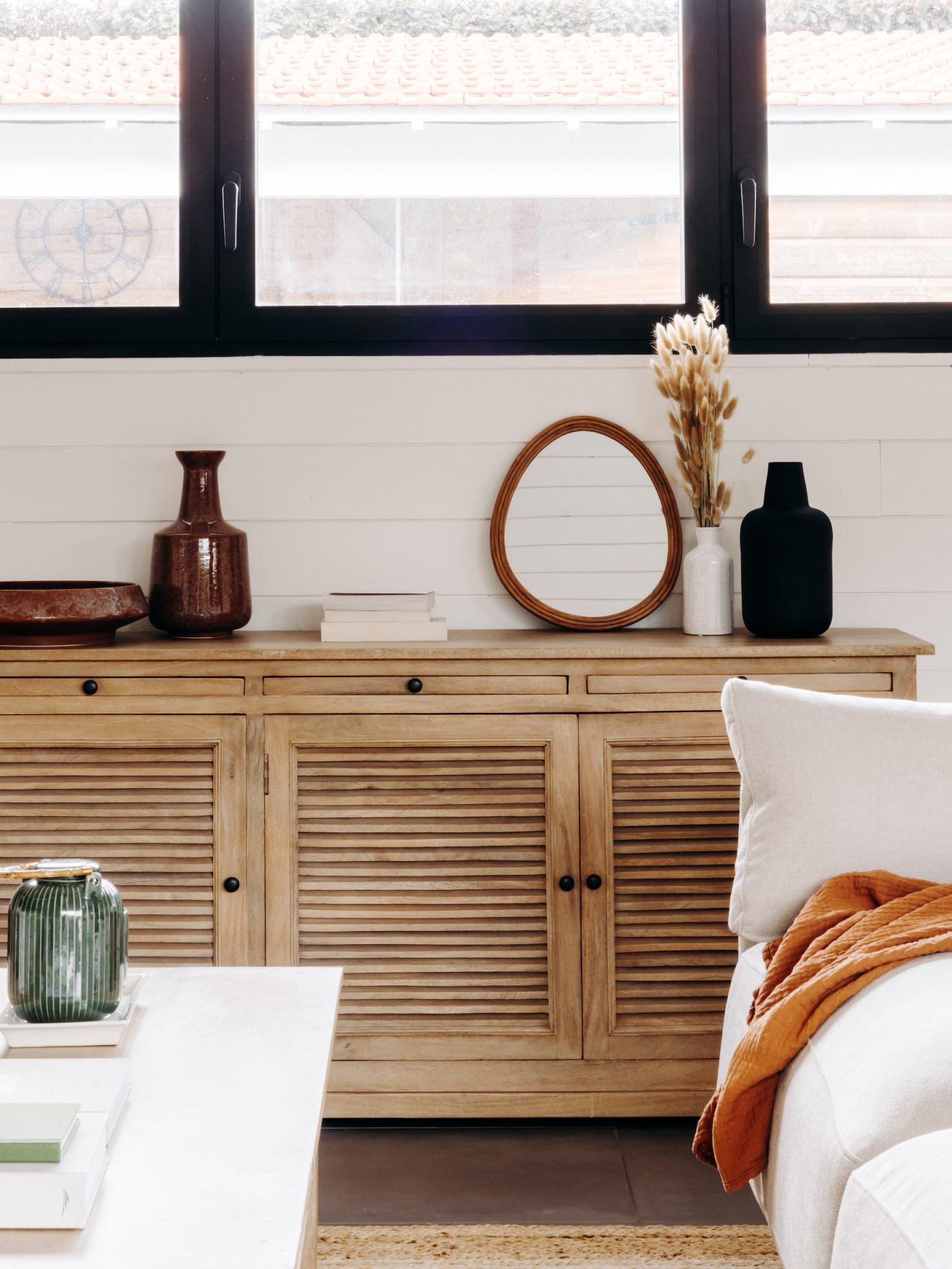 A cozy living room features a wooden sideboard with louvered doors. Above it, there’s an oval mirror, vases, dried plants, and stacked books. A white sofa with an orange throw is in the foreground. Large windows above the sideboard let in natural light.