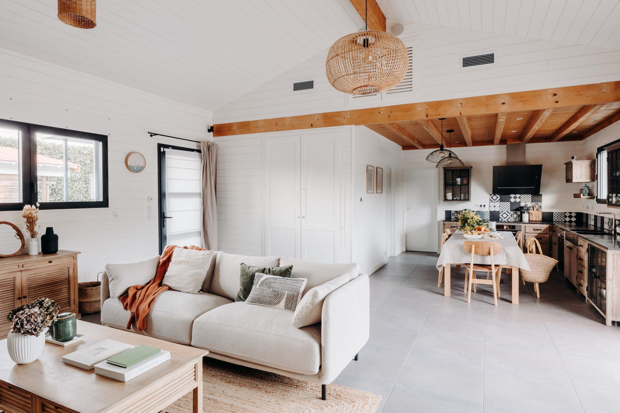 A cozy living room and kitchen corner with a modern rustic design. The living room features a white sofa with cushions, a wooden coffee table, and decorative accents. The kitchen has wooden cabinets, a dining table with chairs, and a pendant light hanging from a ceiling with wooden beams.