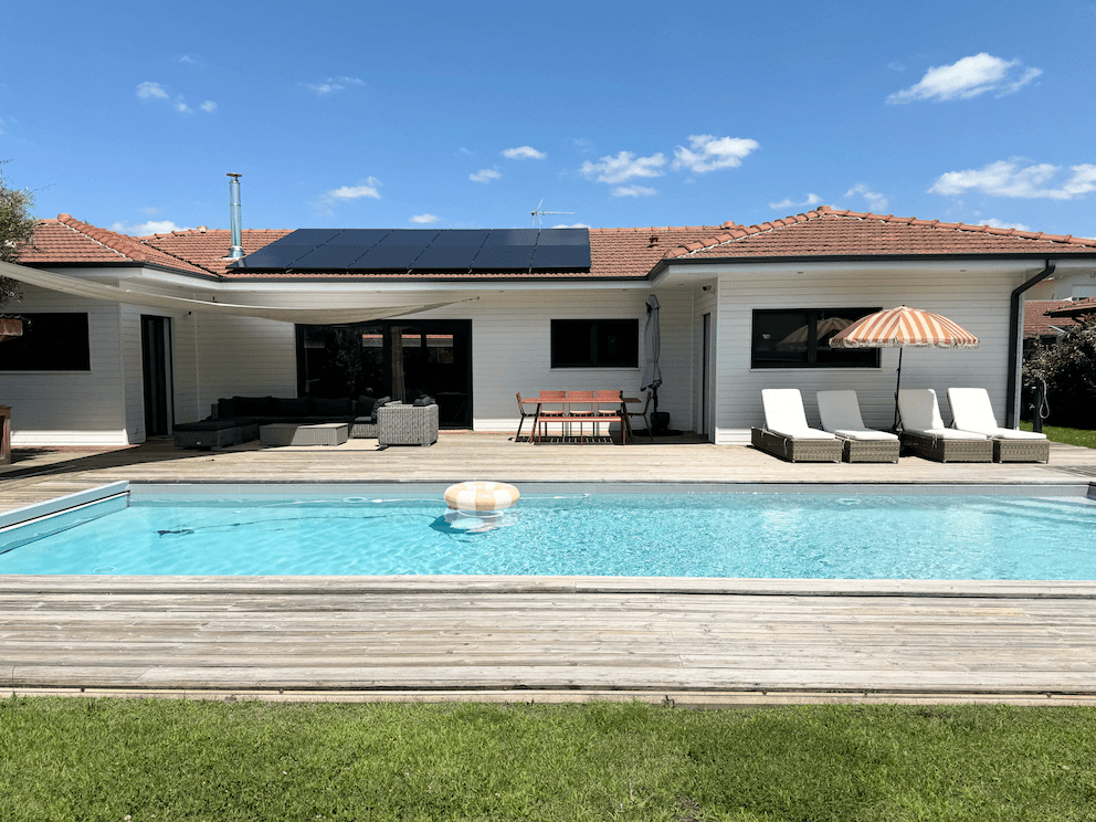 Une maison moderne de plain-pied avec des panneaux solaires sur le toit, un patio avec mobilier de jardin et une piscine au premier plan. La piscine est équipée d'une chaise longue, d'un parasol et d'un anneau gonflable. Le temps est ensoleillé avec un ciel bleu clair.