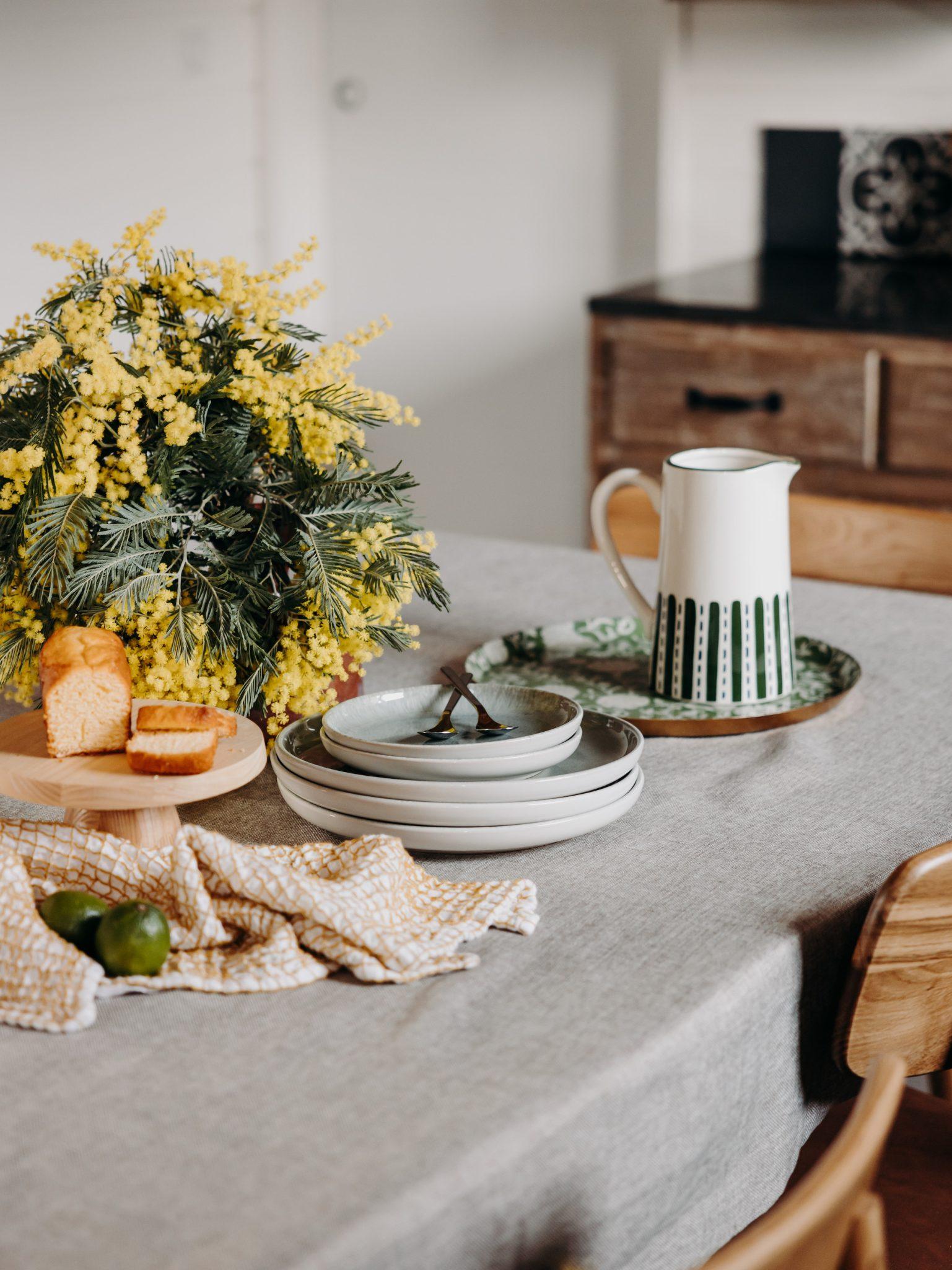 Une table de cuisine dressée sur un thème rustique à la Villa Sixtine, villa à louer pour les vacances au Bassin d'Arcachon. Il comprend un bouquet de fleurs jaunes, une assiette de pain de mie, des assiettes blanches empilées avec une cuillère sur le dessus, un pichet décoratif sur un plateau assorti, un tissu blanc et jaune et deux citrons verts frais.