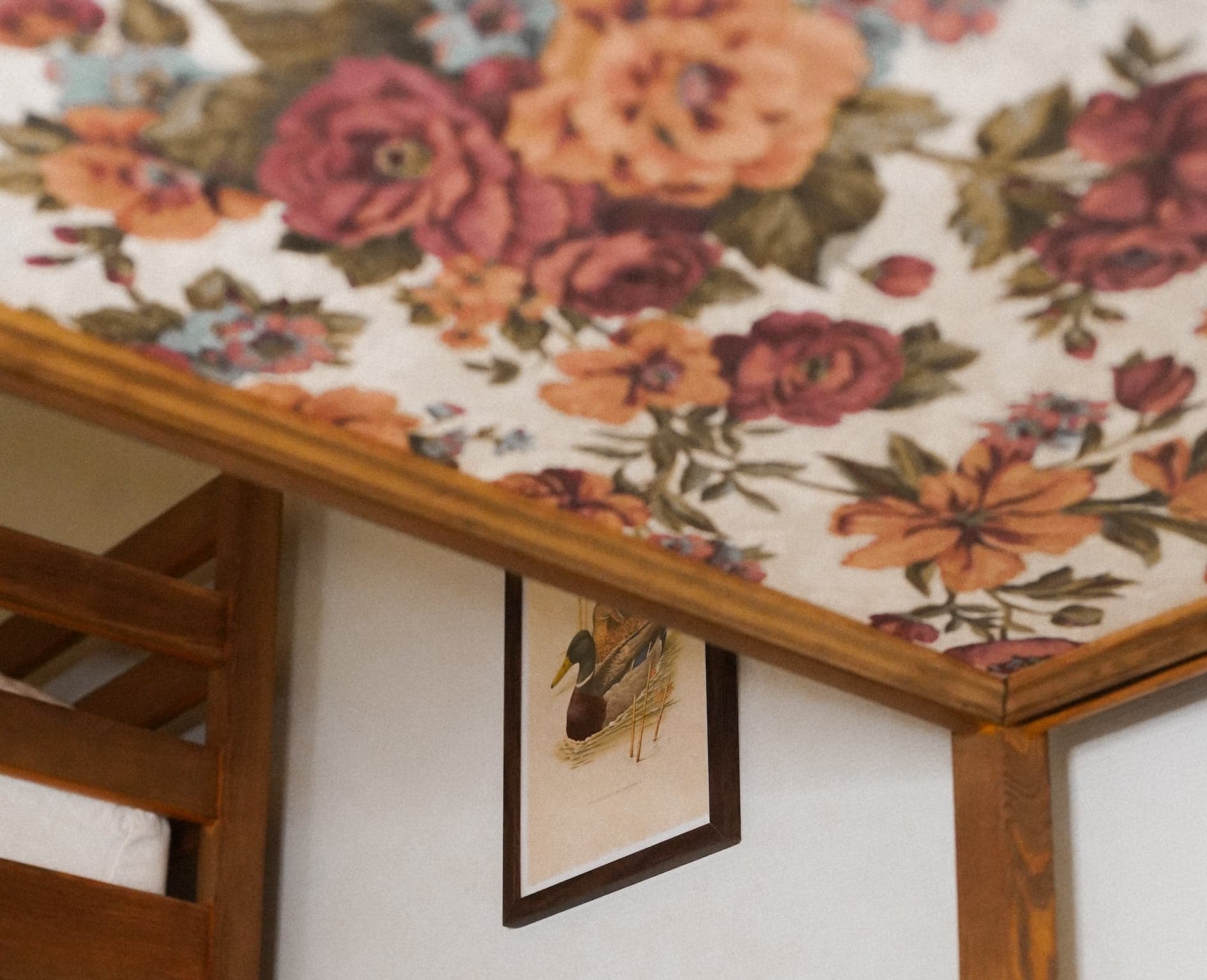 A wooden-framed picture of two ducks, one with a red head and the other with a green head, hanging on a wall beneath a ceiling adorned with vibrant, large floral patterns in red, orange, and green hues. Part of a wooden bunk bed is visible on the left.