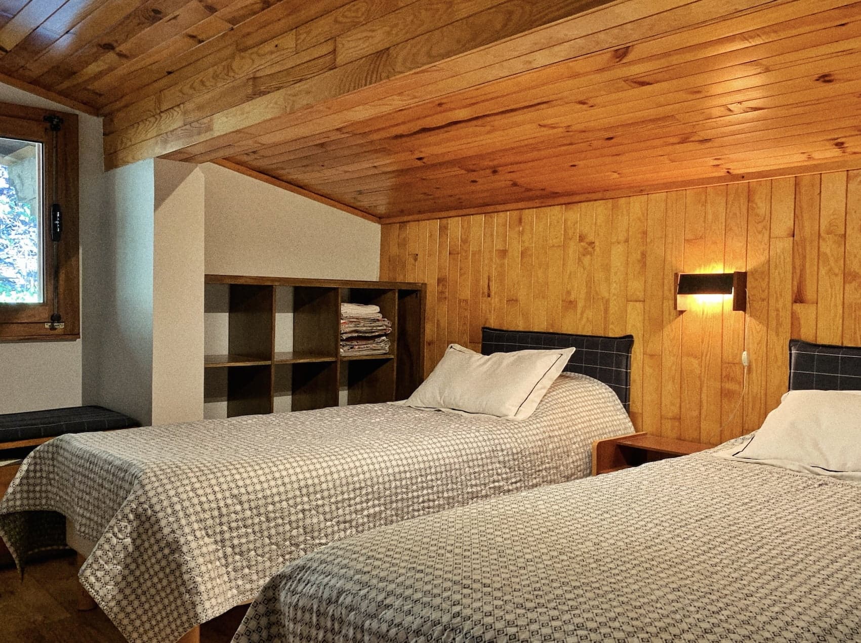 A cozy bedroom with two single beds, each adorned with checkered bedding and pillows. A wooden shelf unit between the beds holds folded blankets. The walls and ceiling are paneled in wood, enhancing the rustic charm. A window and wall lamp provide light.