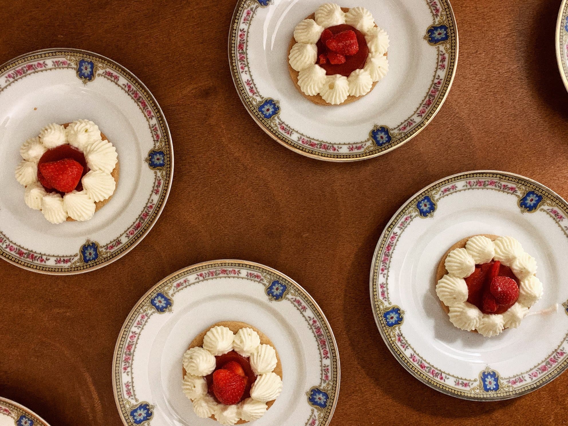 Several ornate porcelain plates are arranged on a wooden table, each holding a small dessert with whipped cream and fresh strawberries garnished on top. The plates feature intricate floral designs with blue and pink accents along their borders.