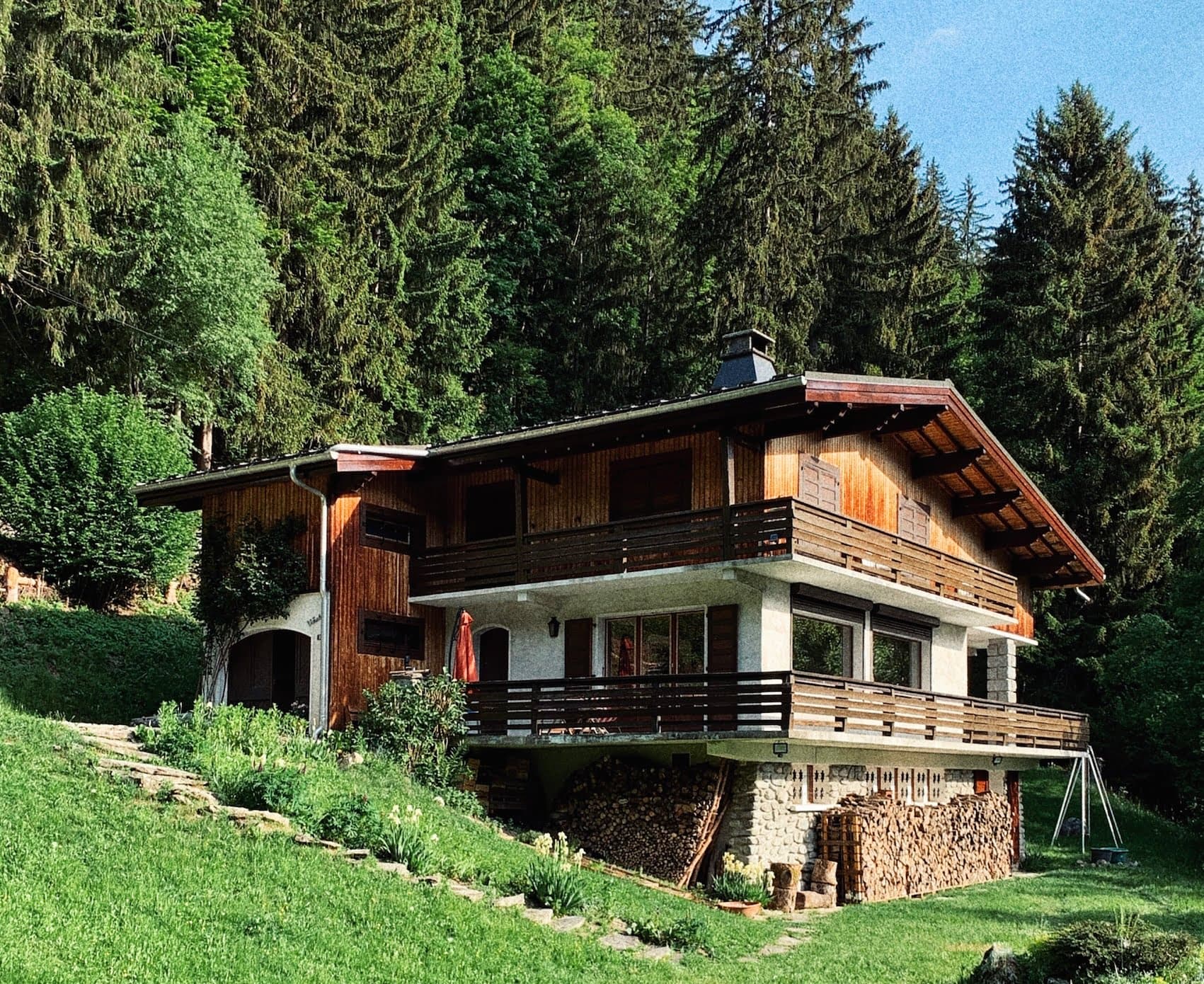 A rustic wooden chalet with a large balcony, situated on a grassy slope and surrounded by dense pine trees. Logs are stacked beneath the balcony, and a swing set is placed on the green lawn. The scene suggests a serene, forested mountain retreat.