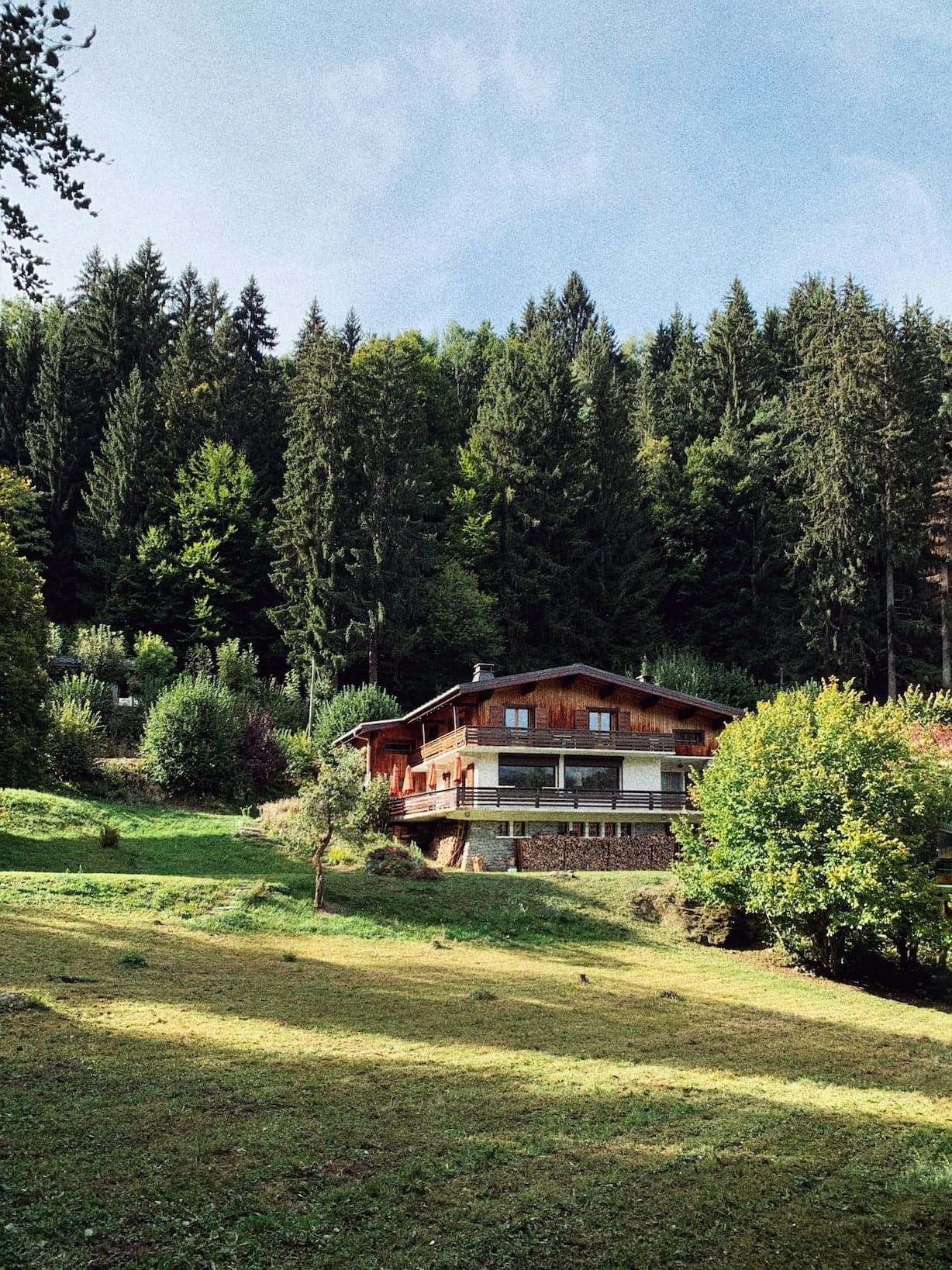 A wooden cabin with a stone foundation is set amidst a grassy area, surrounded by lush green trees. The sun casts dappled shadows on the lawn, and the sky is clear with a hint of scattered clouds. The scene exudes tranquility and natural beauty.