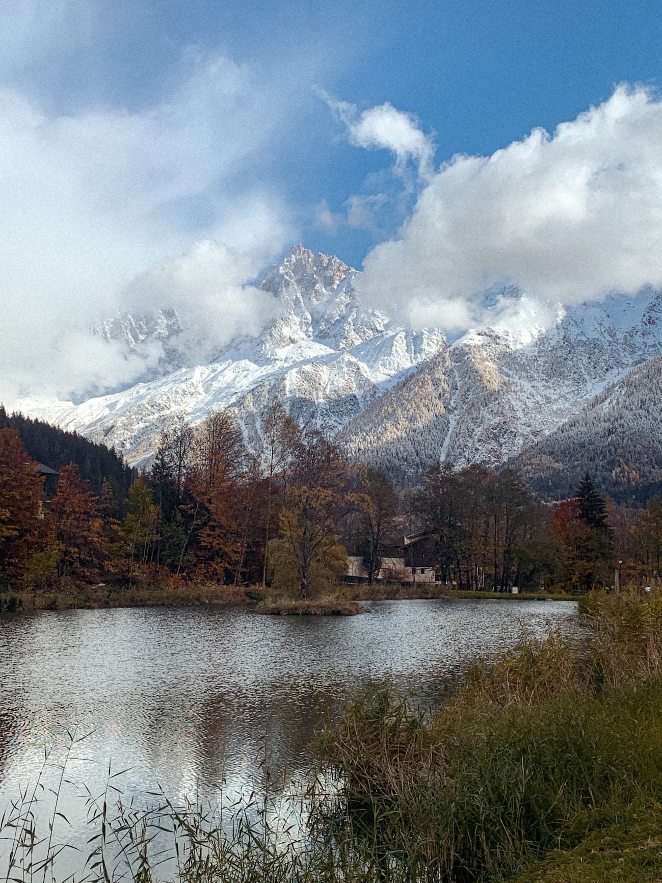 Un lac serein entouré d'arbres aux couleurs automnales reflète le ciel bleu clair, tandis que des montagnes enneigées parsemées de nuages se dressent en arrière-plan. Des roseaux verts luxuriants bordent le bord du lac, ajoutant de la profondeur au paysage tranquille.