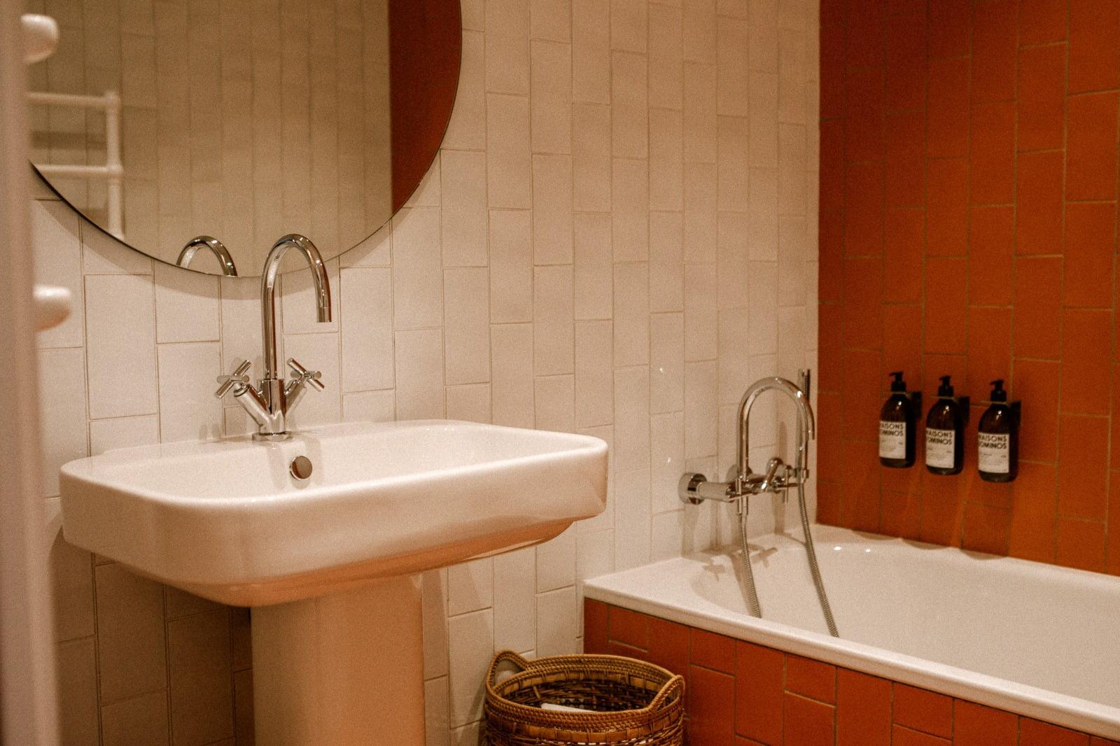 A bathroom with a rectangular white sink featuring a sleek chrome faucet and a round mirror above it, next to a white and brown tiled Domino Deux bathtub with a chrome faucet. There are three bottles mounted on the wall above the bathtub and a wicker basket on the floor.