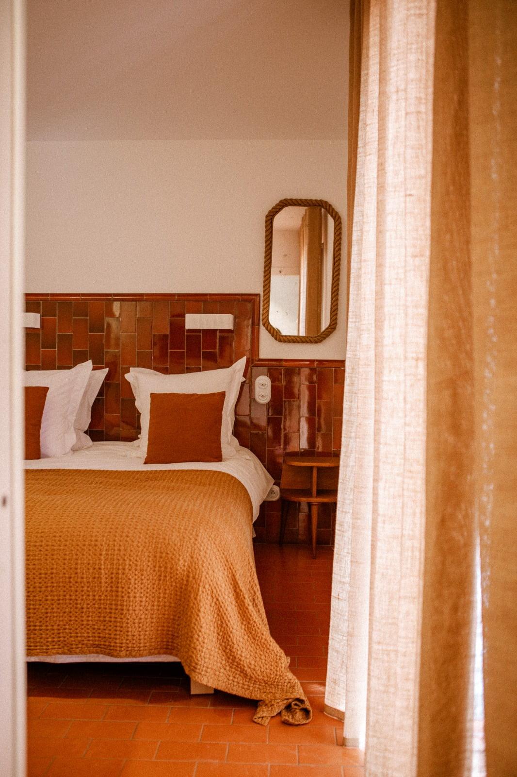 A cozy bedroom with a double bed against a reddish-tiled wall. The bed is adorned with white and brown-orange pillows, along with a matching brown-orange blanket from the Domino Deux collection. A small wooden side table and a mirror are visible in the corner. Light filters through beige curtains.