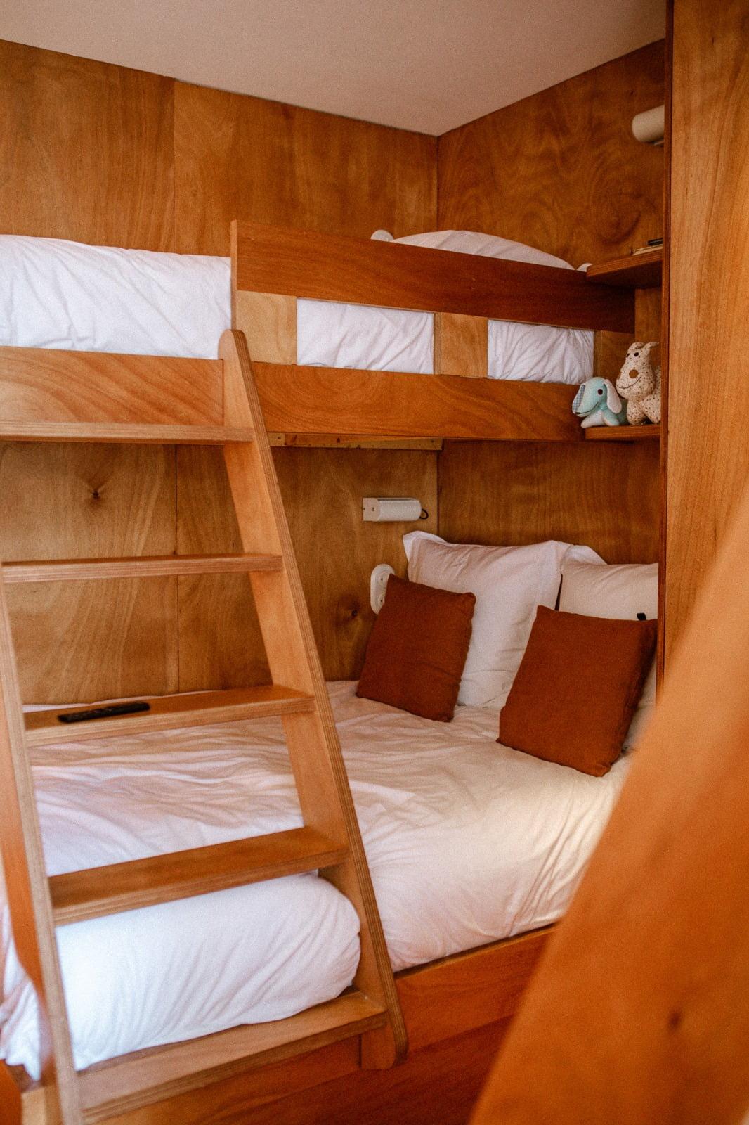 A cozy room with wooden Domino Deux bunk beds. The lower bed is neatly made with white sheets and brown pillows, while the upper bed features a small rabbit and an elephant stuffed animal on the shelf. A wooden ladder provides access to the upper bed, adding to the warm and welcoming atmosphere of the room.