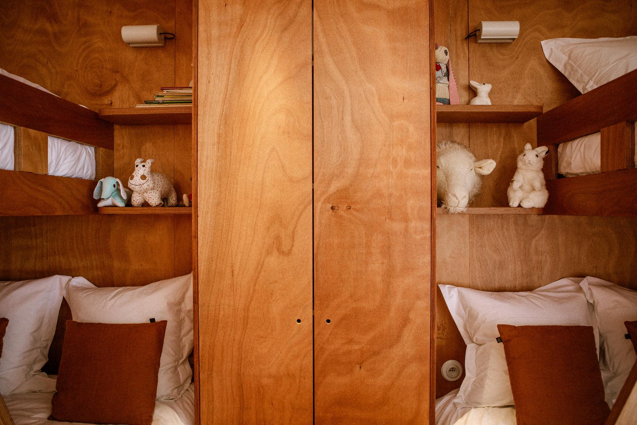 A cozy wooden bunk bed with shelves holding various stuffed animals, including elephants, a bear, a sheep, and a llama. The lower beds are made with white sheets and adorned with brown pillows. Two wall lamps provide the lighting.