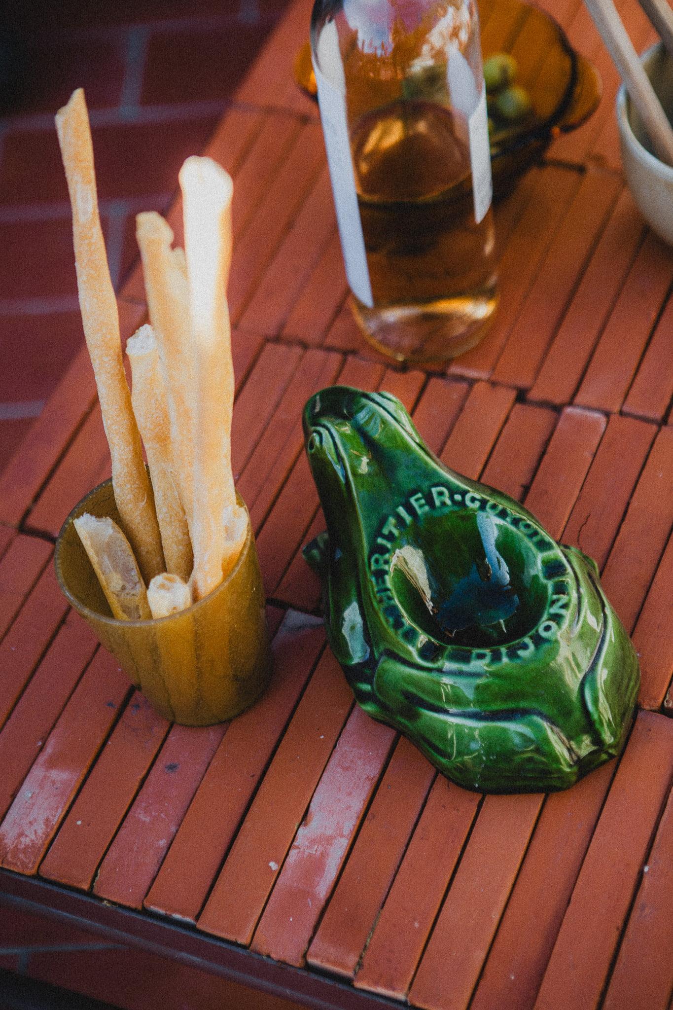 An outdoor table features a cup of breadsticks, a bottle of wine, and a green ceramic ashtray in the shape of a frog with the inscription 'GRAND PRIX' on it. The table has a rustic and lively appearance, set against a backdrop suggesting a casual gathering.